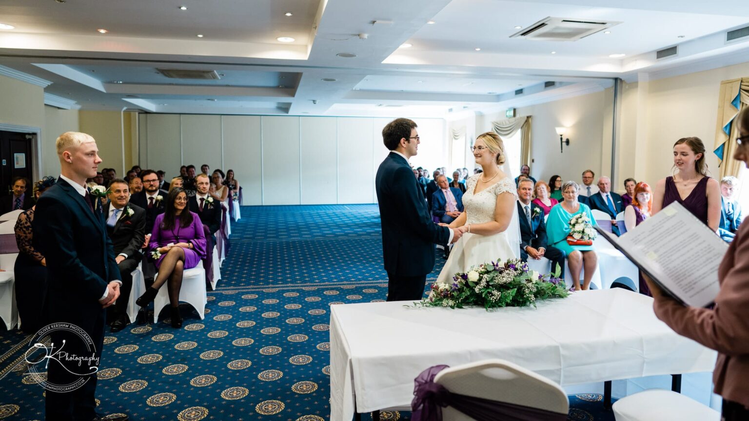Yew Lodge Hotel Wedding Photography Bride and groom exchanging vows at their wedding ceremony in a hotel function room, with guests seated and watching.
