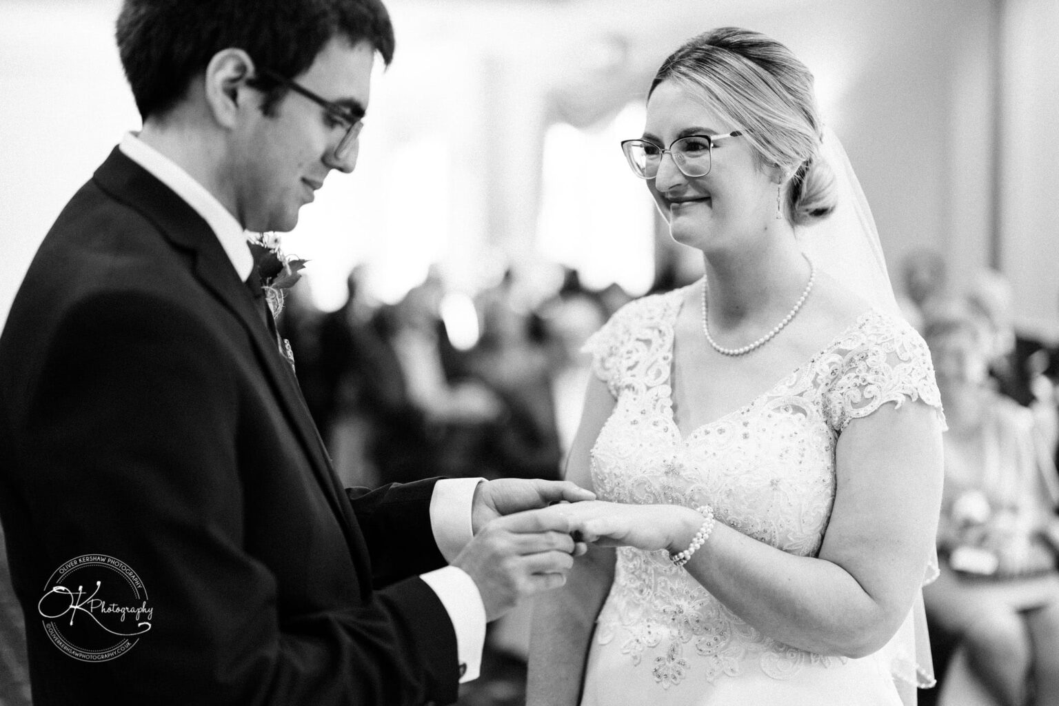 Yew Lodge Hotel Wedding Photography Groom places ring on bride's finger during wedding ceremony, both smiling, black and white photo.