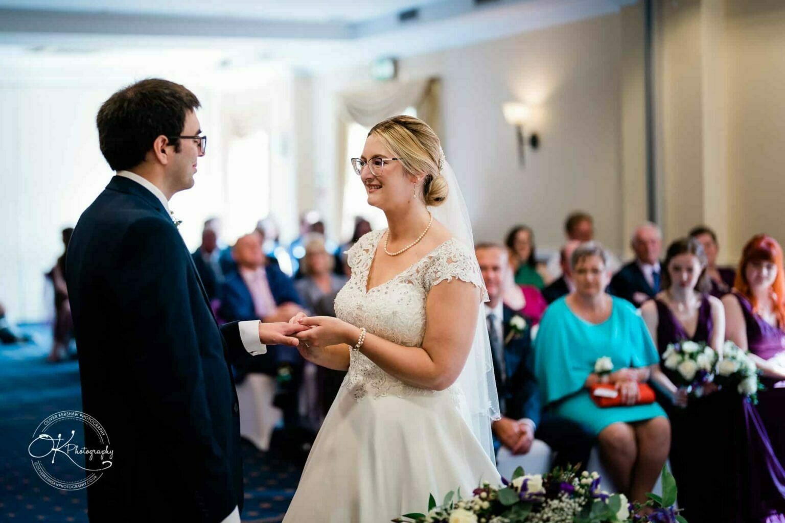Yew Lodge Hotel Wedding Photography Bride and groom exchanging rings during their wedding ceremony at Yew Lodge Hotel, with guests seated in the background.