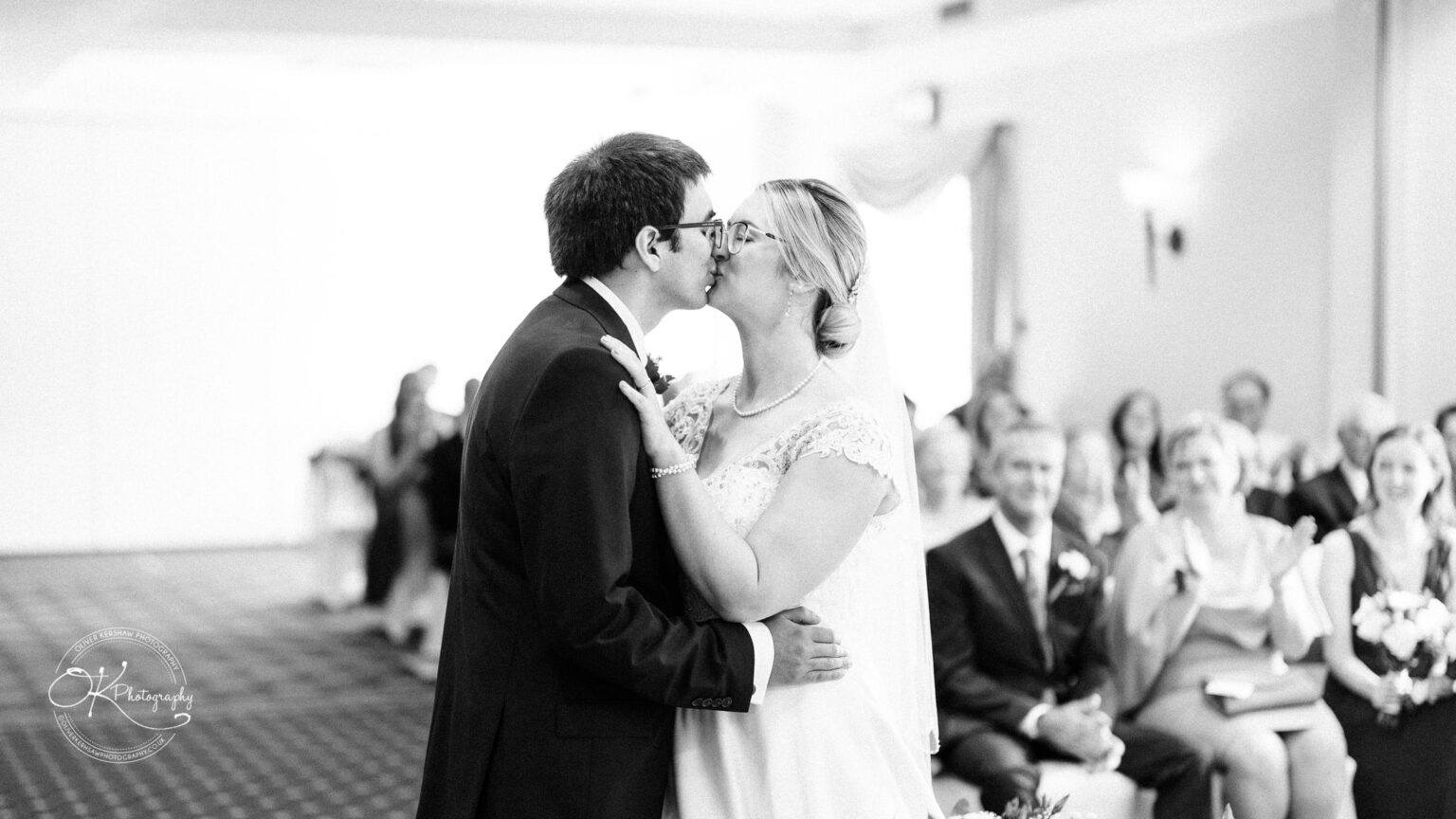 Yew Lodge Hotel Wedding Photography A newlywed couple shares a kiss during their wedding ceremony, surrounded by guests clapping and smiling in the audience at the Yew Lodge Hotel.