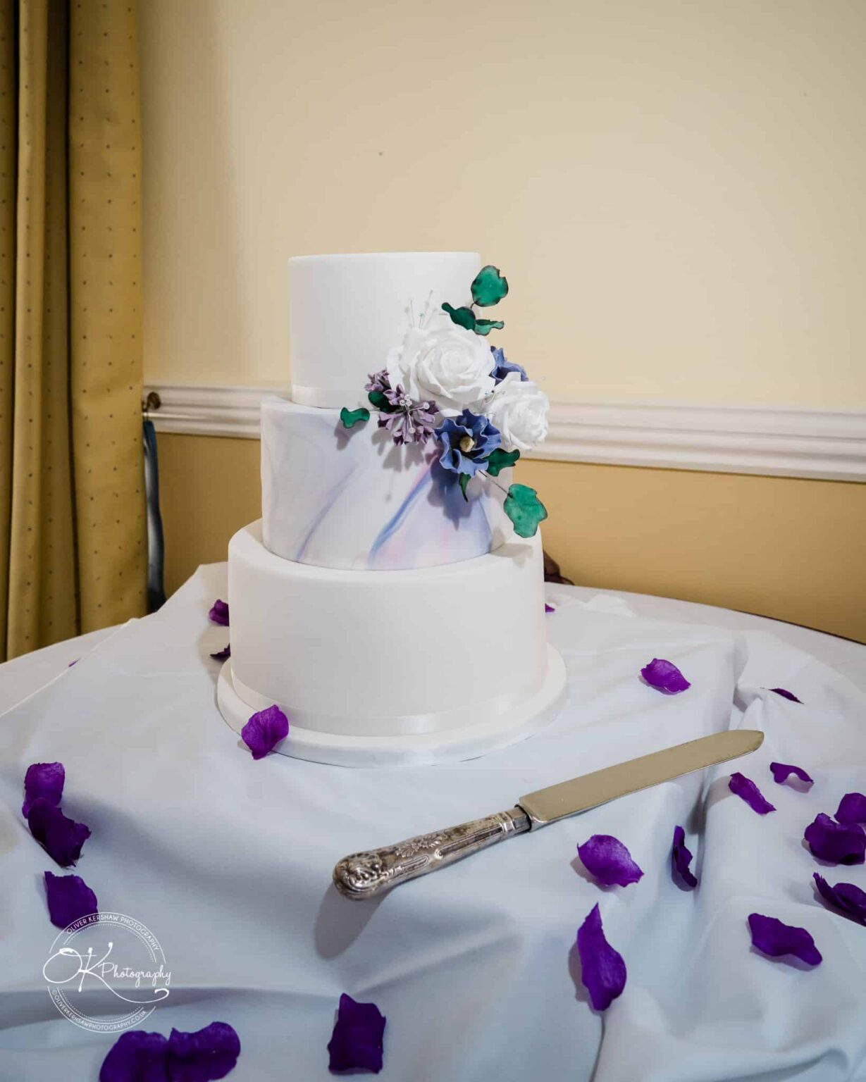 Yew Lodge Hotel Wedding Photography A three-tiered wedding cake with white icing, floral decorations, and purple petals scattered around, with a cake knife placed beside it.