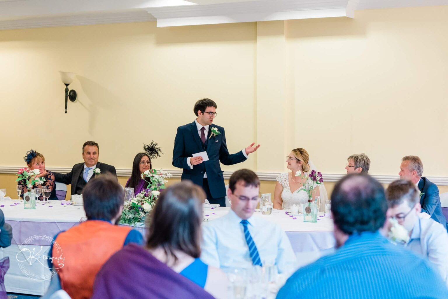 Yew Lodge Hotel Wedding Photography A person in a suit giving a speech at a wedding reception with the bride, groom, and guests seated at a table.