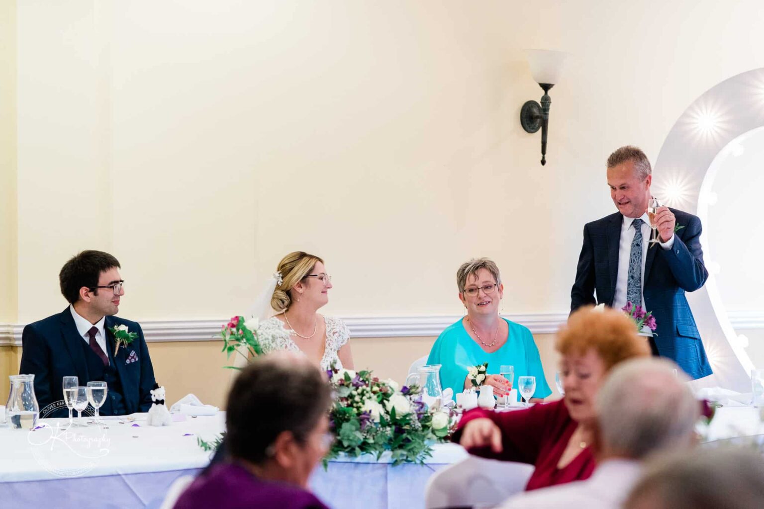 Yew Lodge Hotel Wedding Photography Bride and groom seated at a wedding reception table with family members, while a man stands raising a toast.