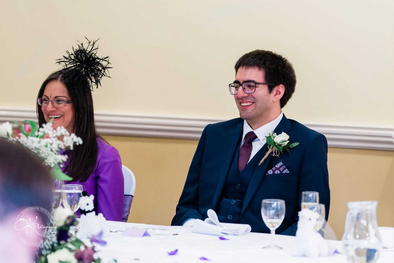 Yew Lodge Hotel Wedding Photography People seated at a table during a wedding, dressed in formal attire, and smiling; the woman is wearing a fascinator.