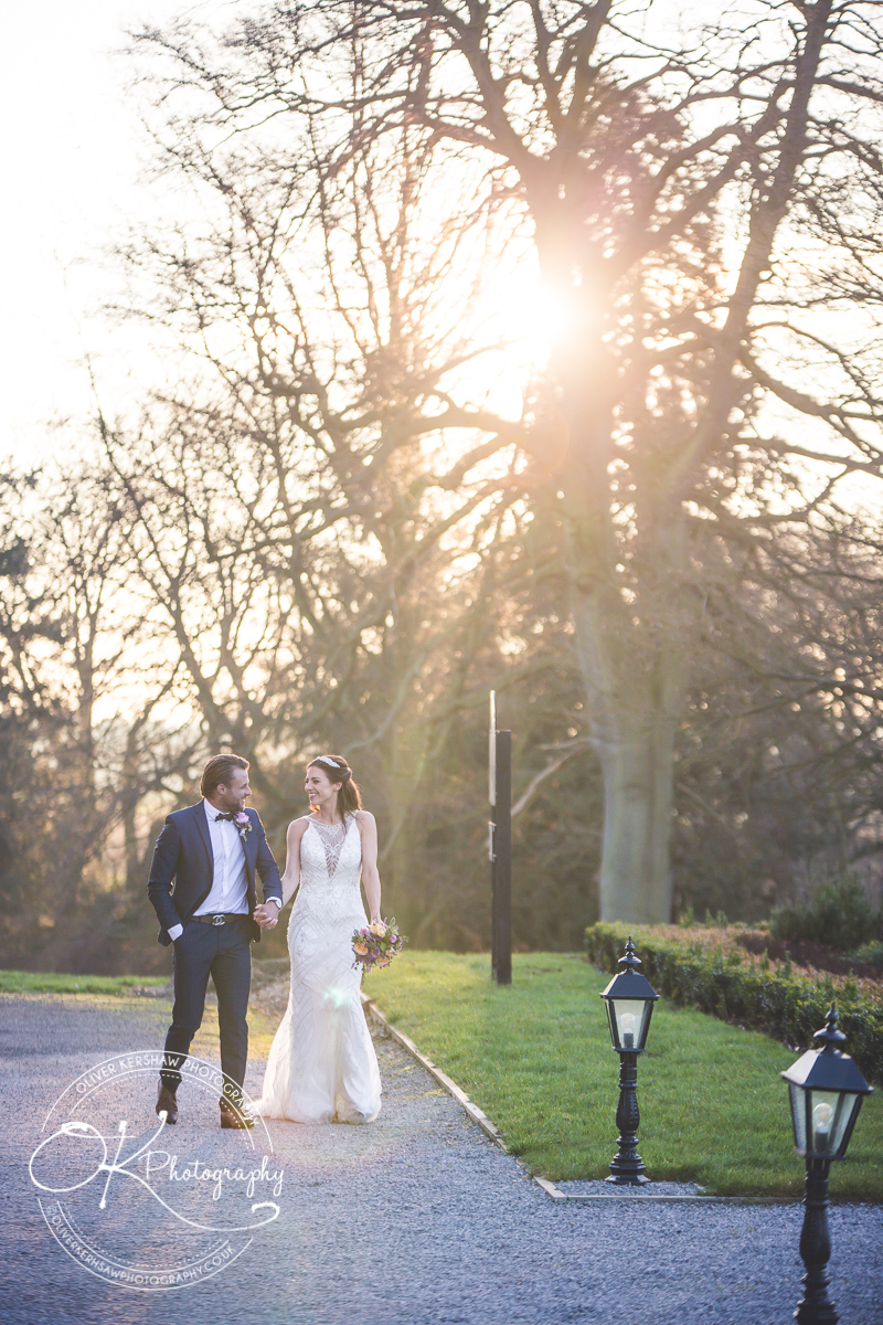 A bride and groom walking hand-in-hand outside, with the bride in a white wedding dress holding a bouquet and the groom in a dark suit with a bow tie, in front of a sunlit backdrop of trees.