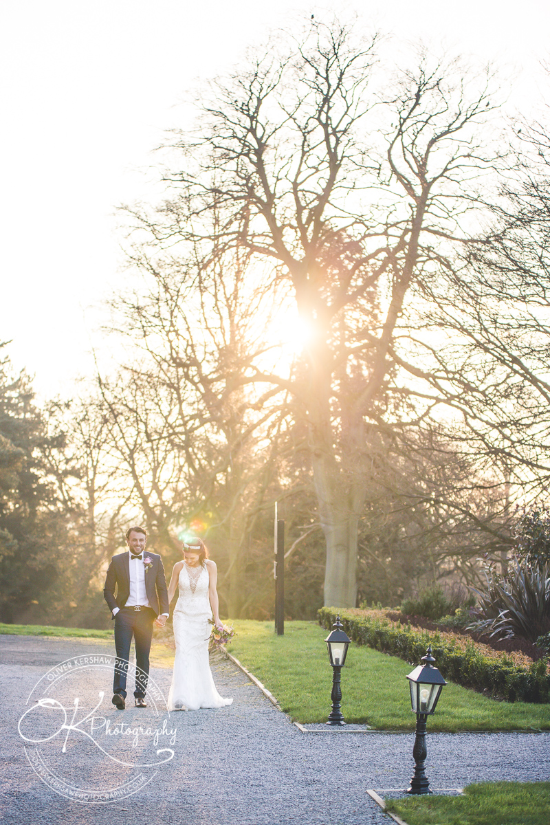 A bride and groom walking hand in hand on a path surrounded by green grass and trees with the sun shining through the branches.