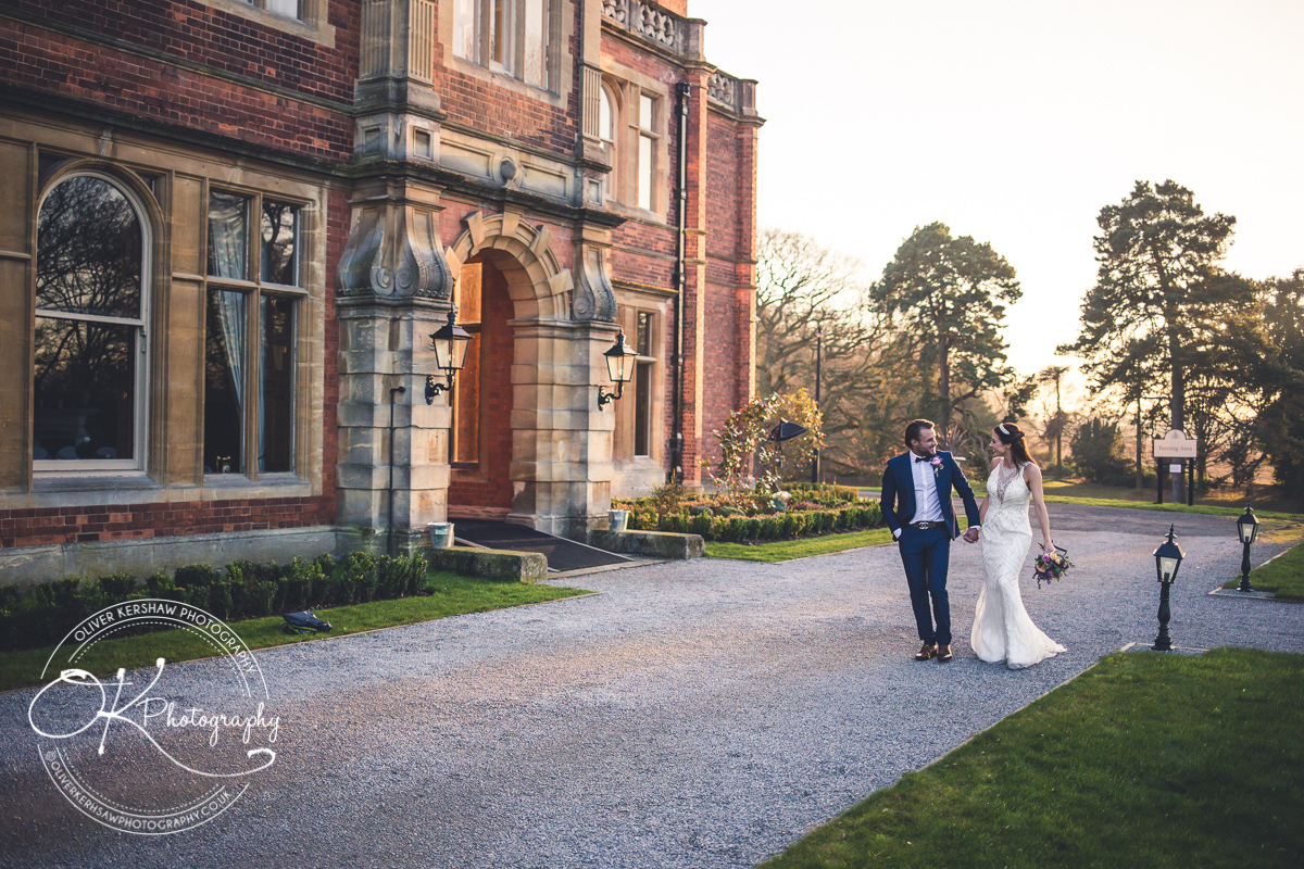Bride and groom walking hand in hand outside a large historic building on a sunny day.