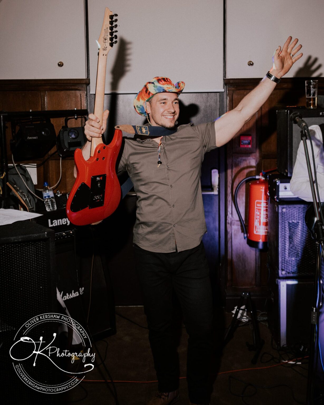 Wedding Photography George Hotel Stamford A man holding a red electric guitar and wearing a colorful hat, raising one arm in the air, standing in a room with musical equipment.