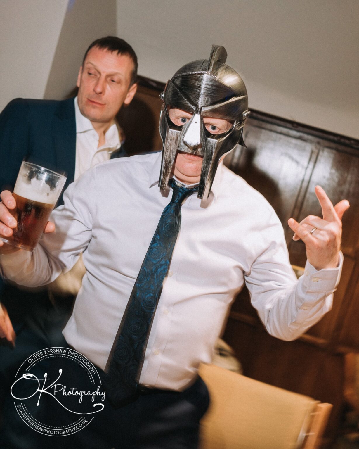 Wedding Photography George Hotel Stamford A man wearing a metallic gladiator helmet and a white dress shirt holds a pint of beer, with another man in a suit standing behind him.