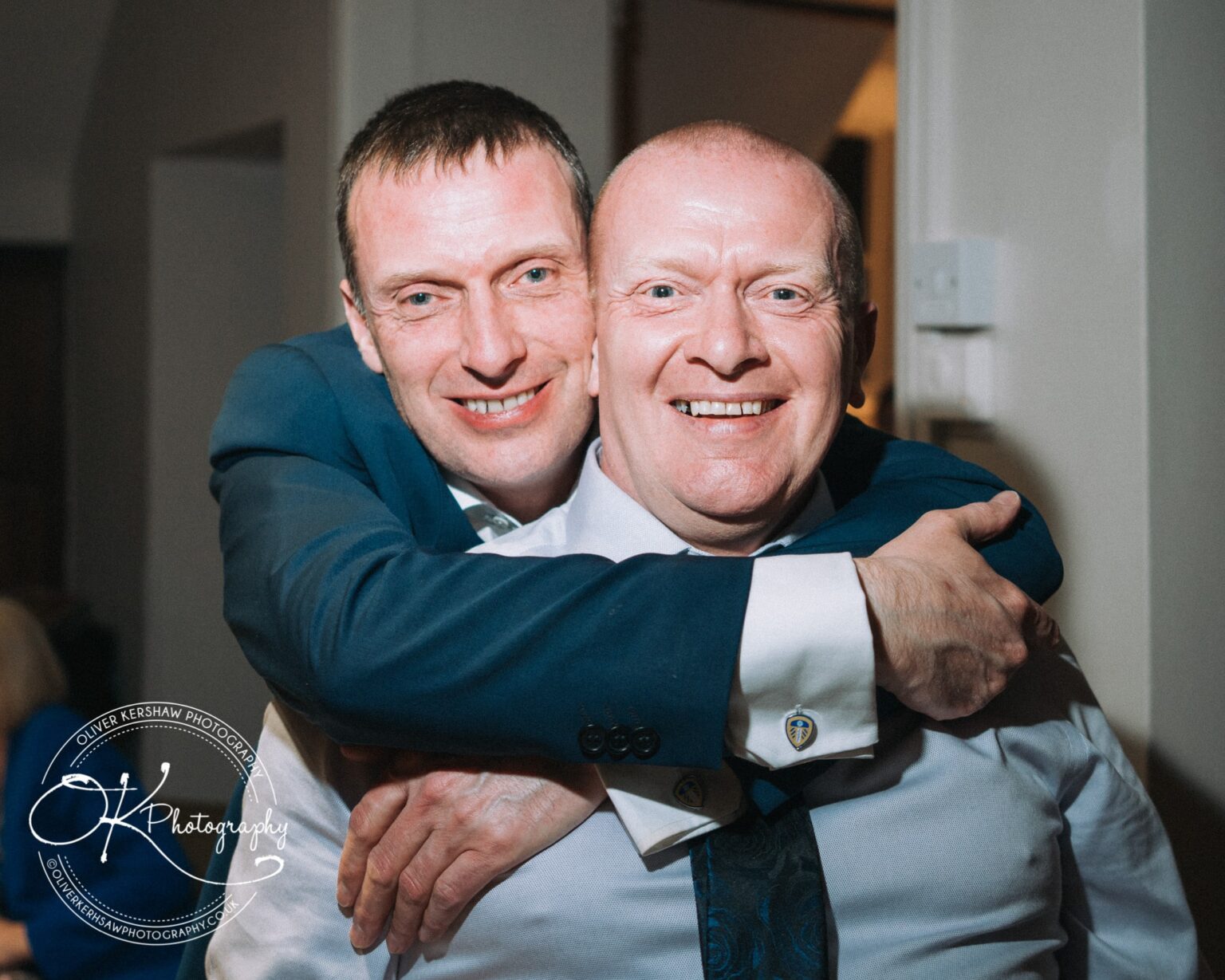Wedding Photography George Hotel Stamford Two smiling men embracing, one in a white shirt and tie, the other in a blue suit jacket, indoors at the George Hotel, Stamford.