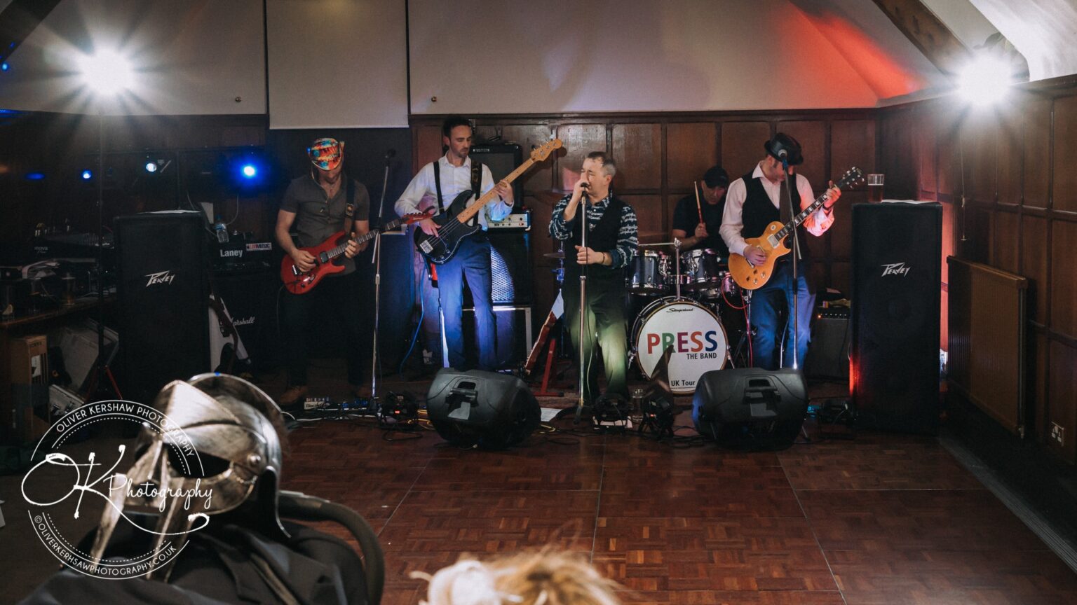 Wedding Photography George Hotel Stamford A band performing live on stage in a room with wooden panelled walls. The drum kit displays a logo "PRESS THE BAND." There is stage lighting and sound equipment around them.