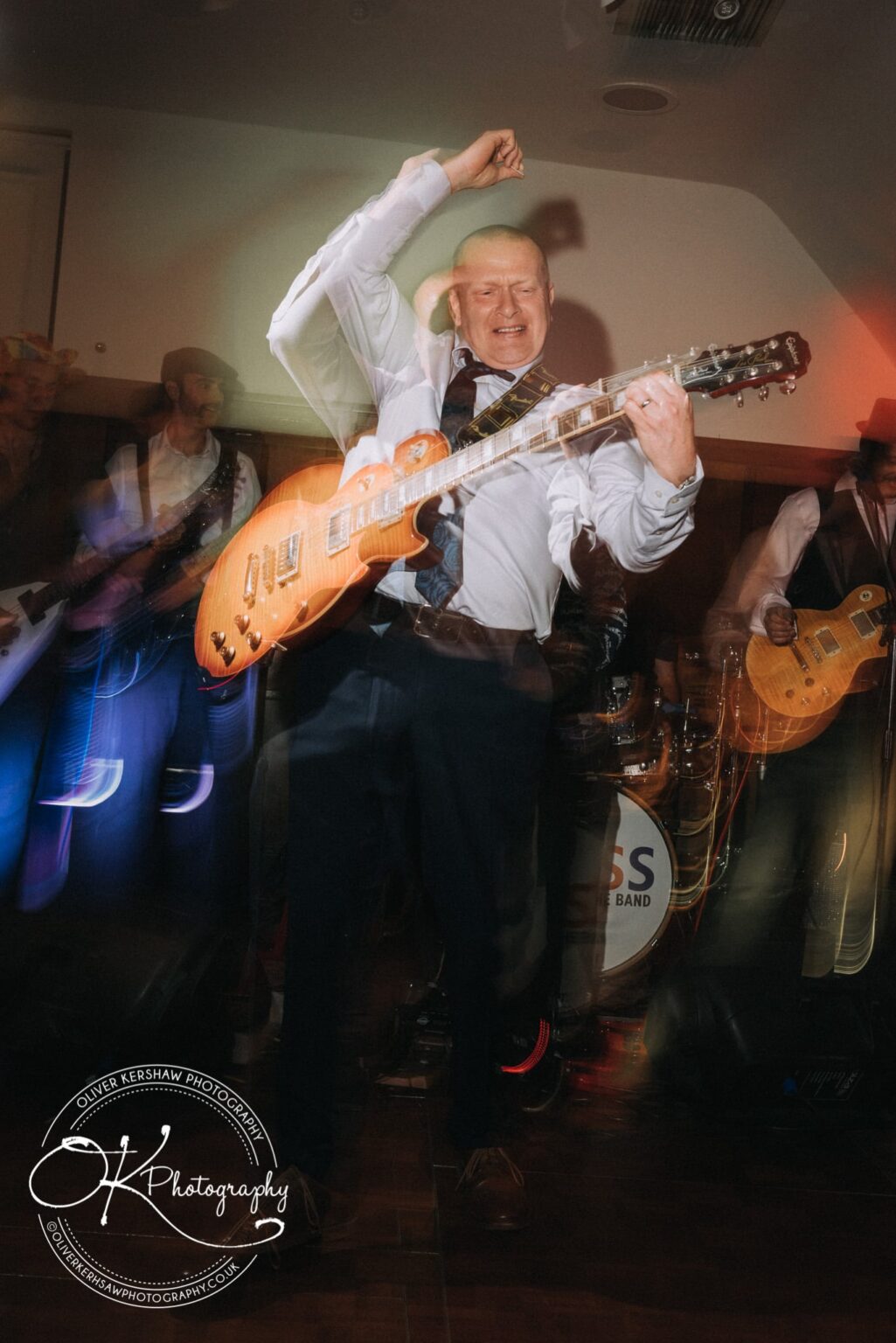 Wedding Photography George Hotel Stamford A man in a white shirt and tie enthusiastically playing a guitar on stage with a band, with blurred motion effects and colourful stage lighting.