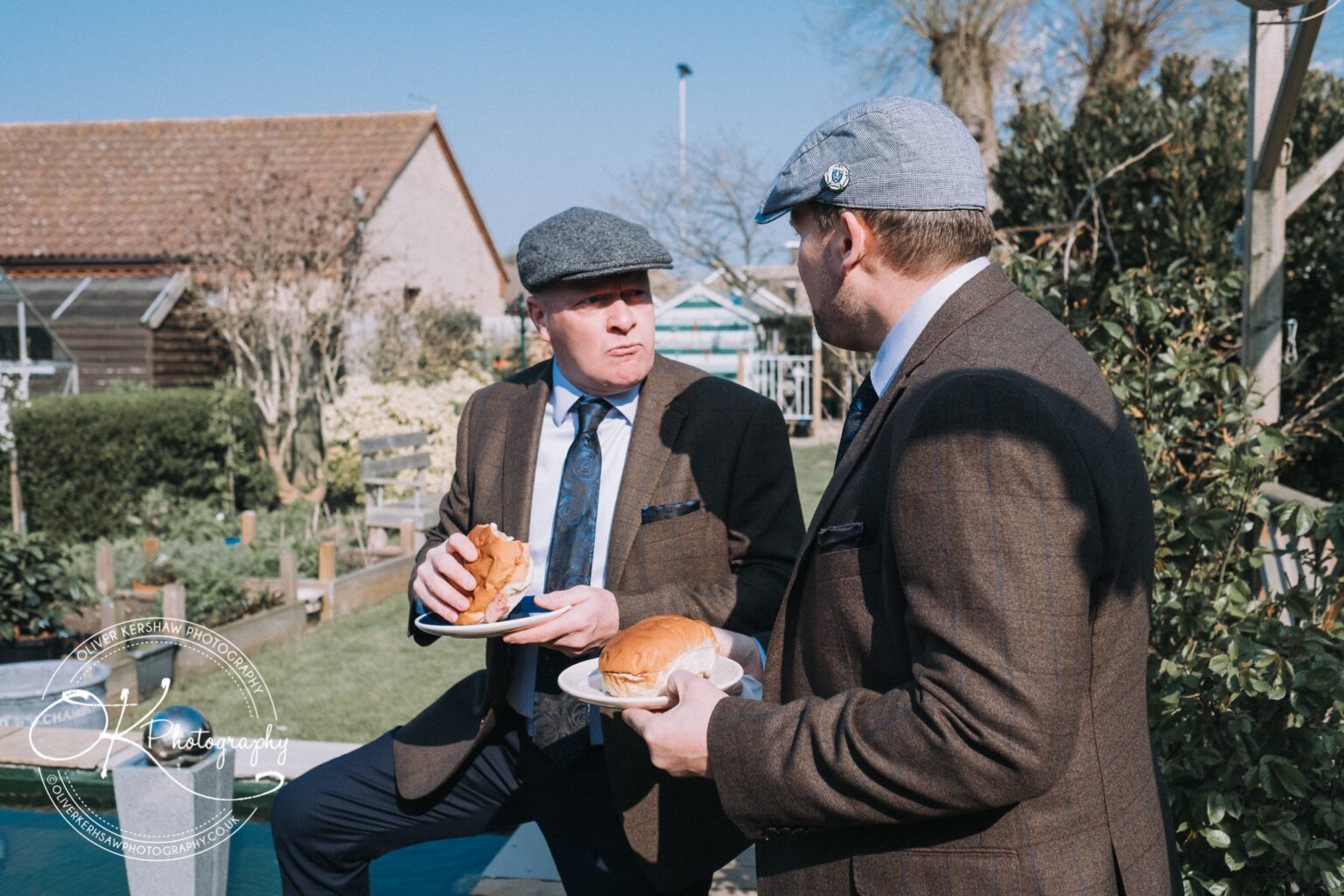 Wedding Photography George Hotel Stamford Two men in suits and flat caps eating sandwiches outside on a sunny day.