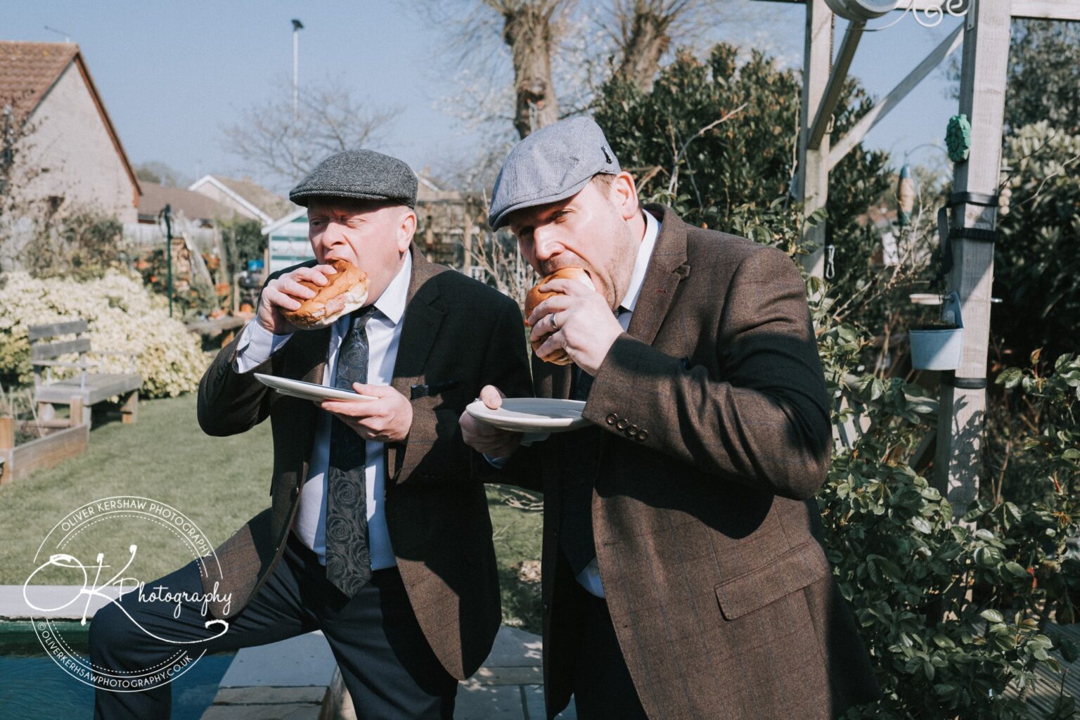 Wedding Photography George Hotel Stamford Two men in suits and flat caps eating sandwiches in a garden.