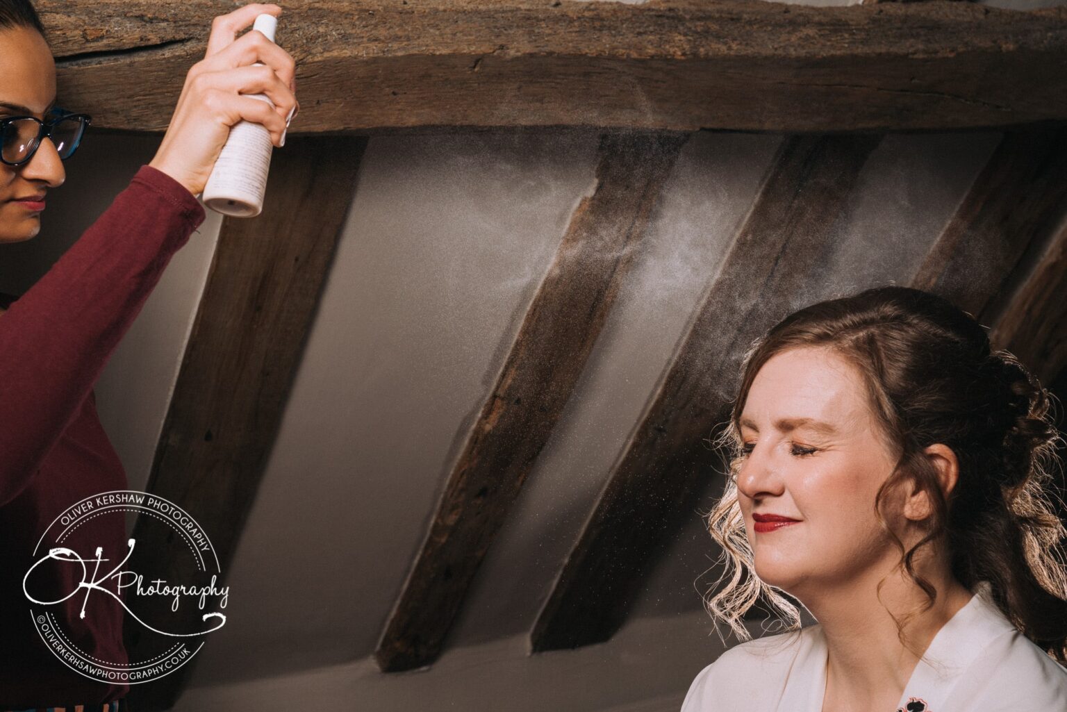 Wedding Photography George Hotel Stamford A woman with closed eyes having hairspray applied in a rustic room.