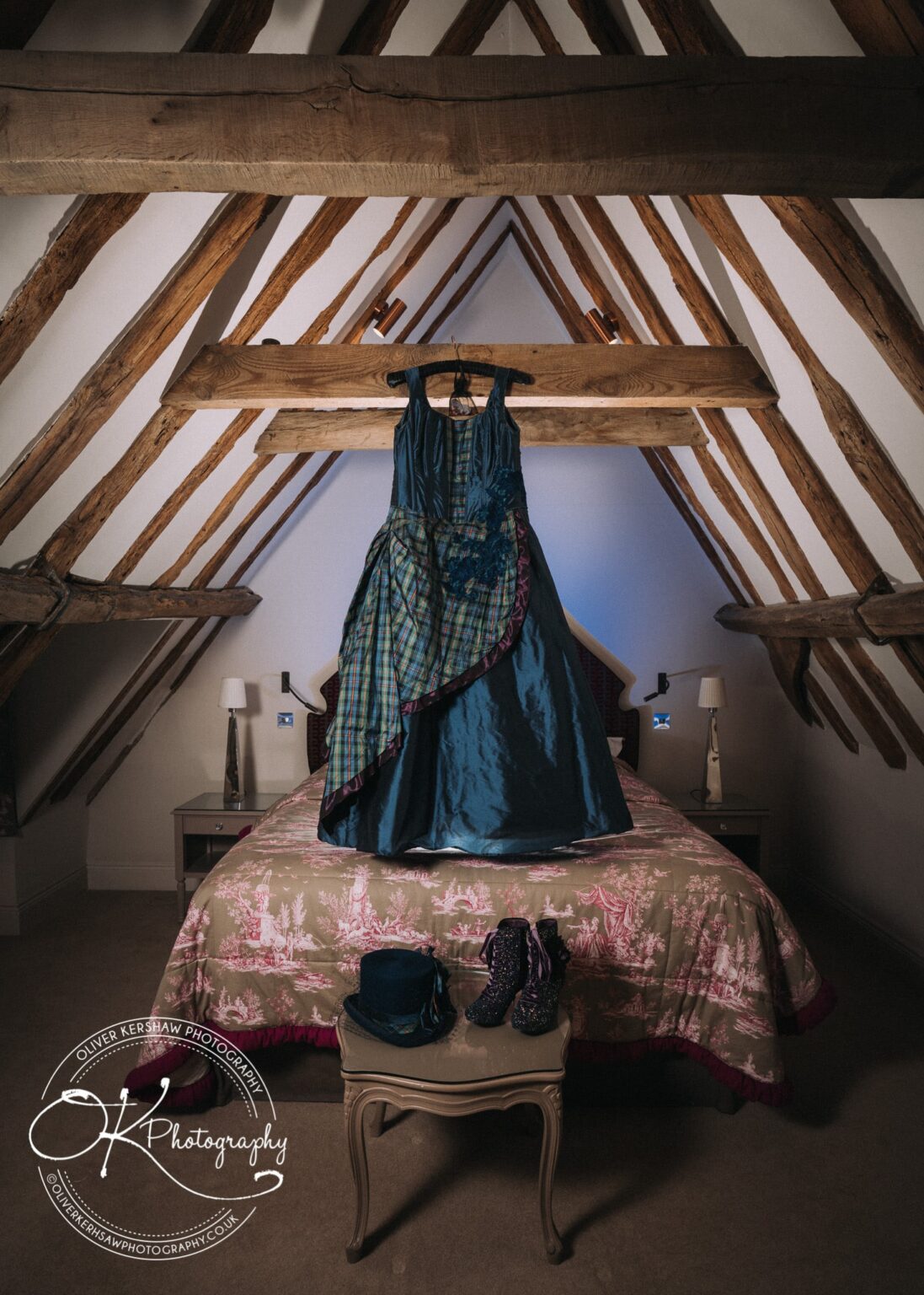 Wedding Photography George Hotel Stamford A blue dress with a green plaid overlay hangs from a wooden beam in an attic room with exposed wooden rafters. Below the dress is a bed with a patterned quilt, and a small table in front holds a blue hat and a pair of sequined boots.