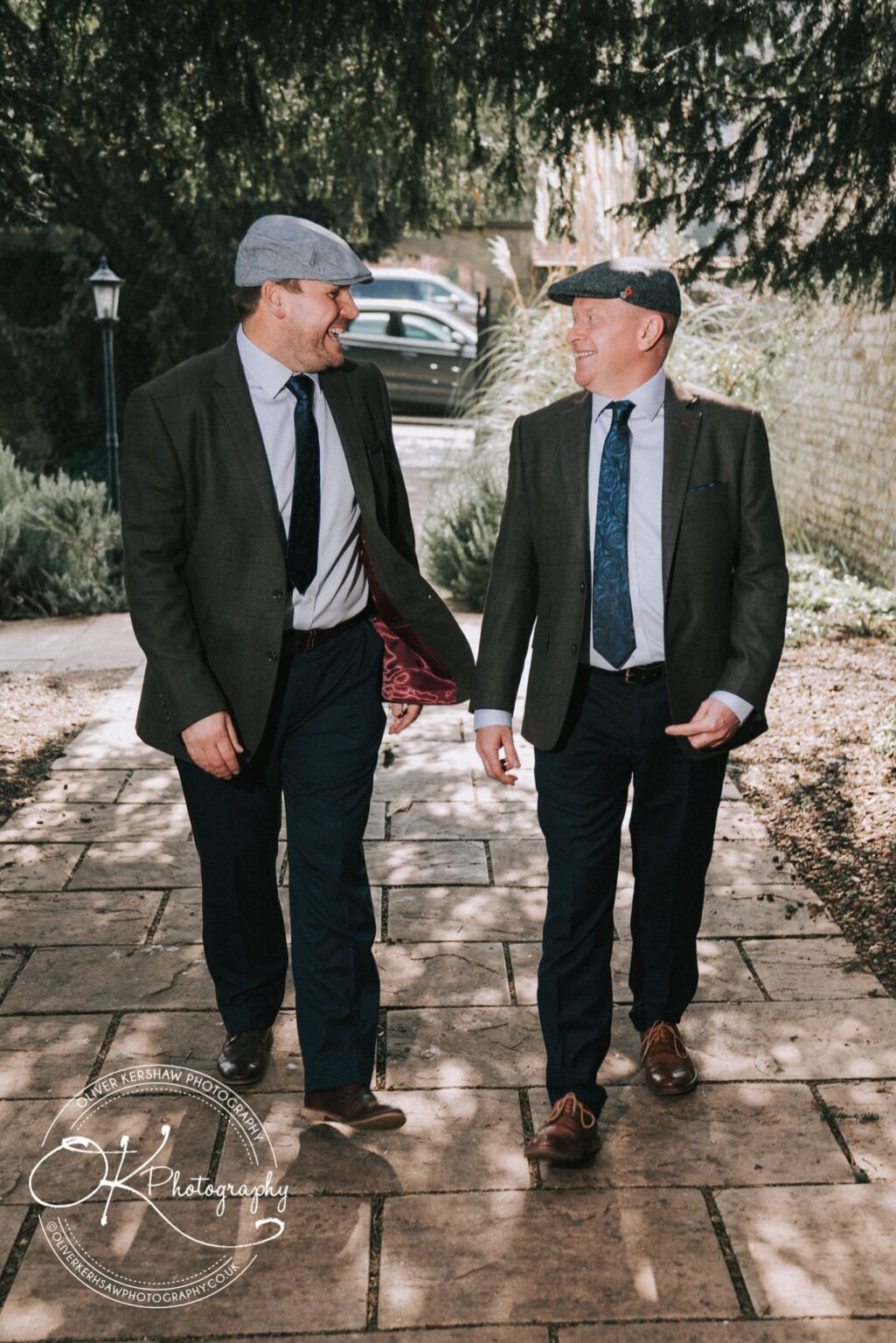 Wedding Photography George Hotel Stamford Two men in suits and flat caps walking on a stone pathway, smiling and talking to each other.