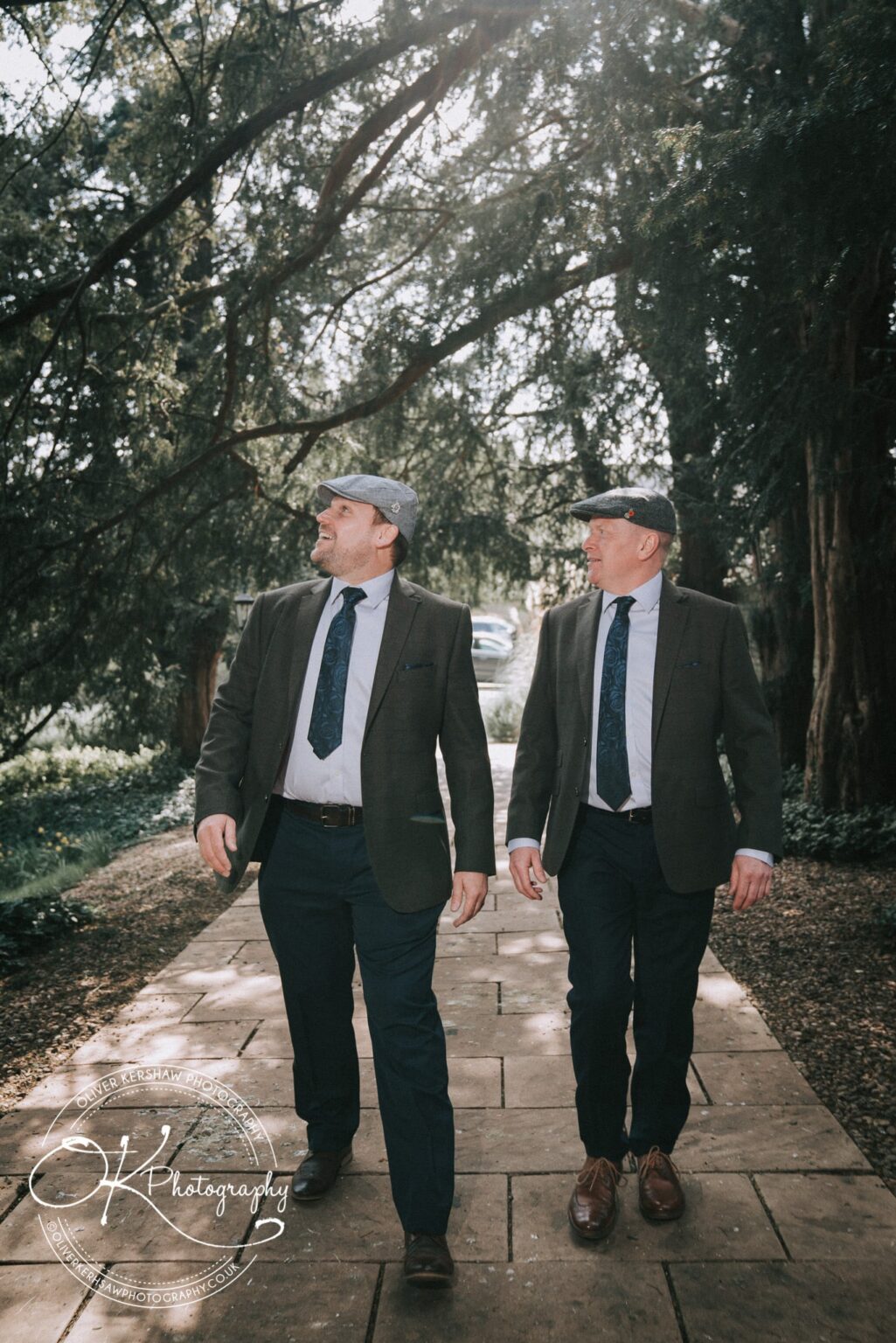 Wedding Photography George Hotel Stamford Two men wearing suits and flat caps walking on a path surrounded by trees and greenery.