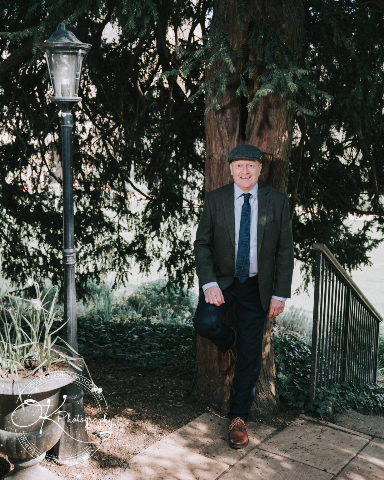 Wedding Photography George Hotel Stamford A man in a suit and flat cap stands outdoors by a tree and an old-fashioned streetlamp, leaning against the tree with one leg bent. A metal railing and garden planter with flowers are nearby.
