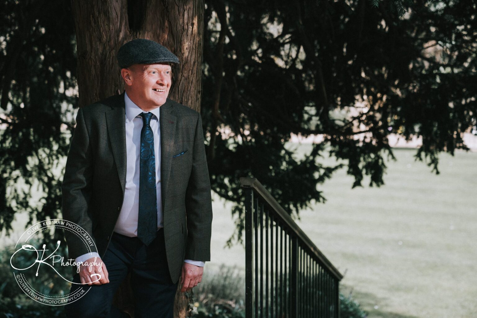 Wedding Photography George Hotel Stamford Man wearing a suit and flat cap, standing outdoors by a tree and iron railing.
