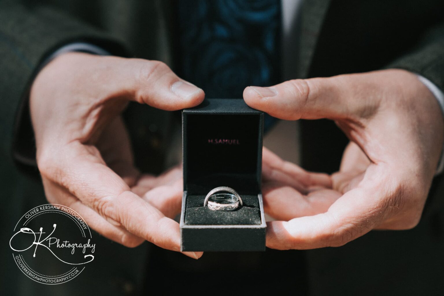 Wedding Photography George Hotel Stamford Hands holding an open black ring box containing a silver ring, with branding from H. Samuel visible inside the box lid. The image has a watermark from Oliver Kershaw Photography.