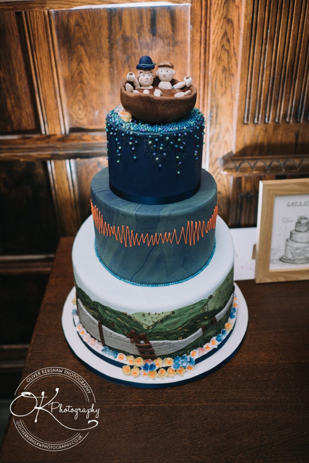 Wedding Photography George Hotel Stamford A three-tiered cake with intricate decorations, featuring a blue top tier with figurines in a hot tub, a middle tier with a green marbled pattern and orange accents, and a bottom tier with a countryside landscape and flowers.
