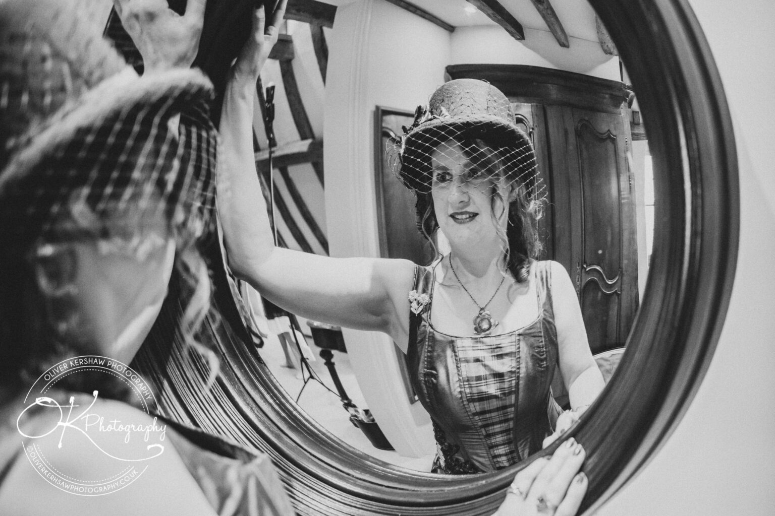 Wedding Photography George Hotel Stamford A woman wearing a vintage outfit and a hat with a net veil admires herself in a round mirror. The setting includes wooden beams and antique furniture.