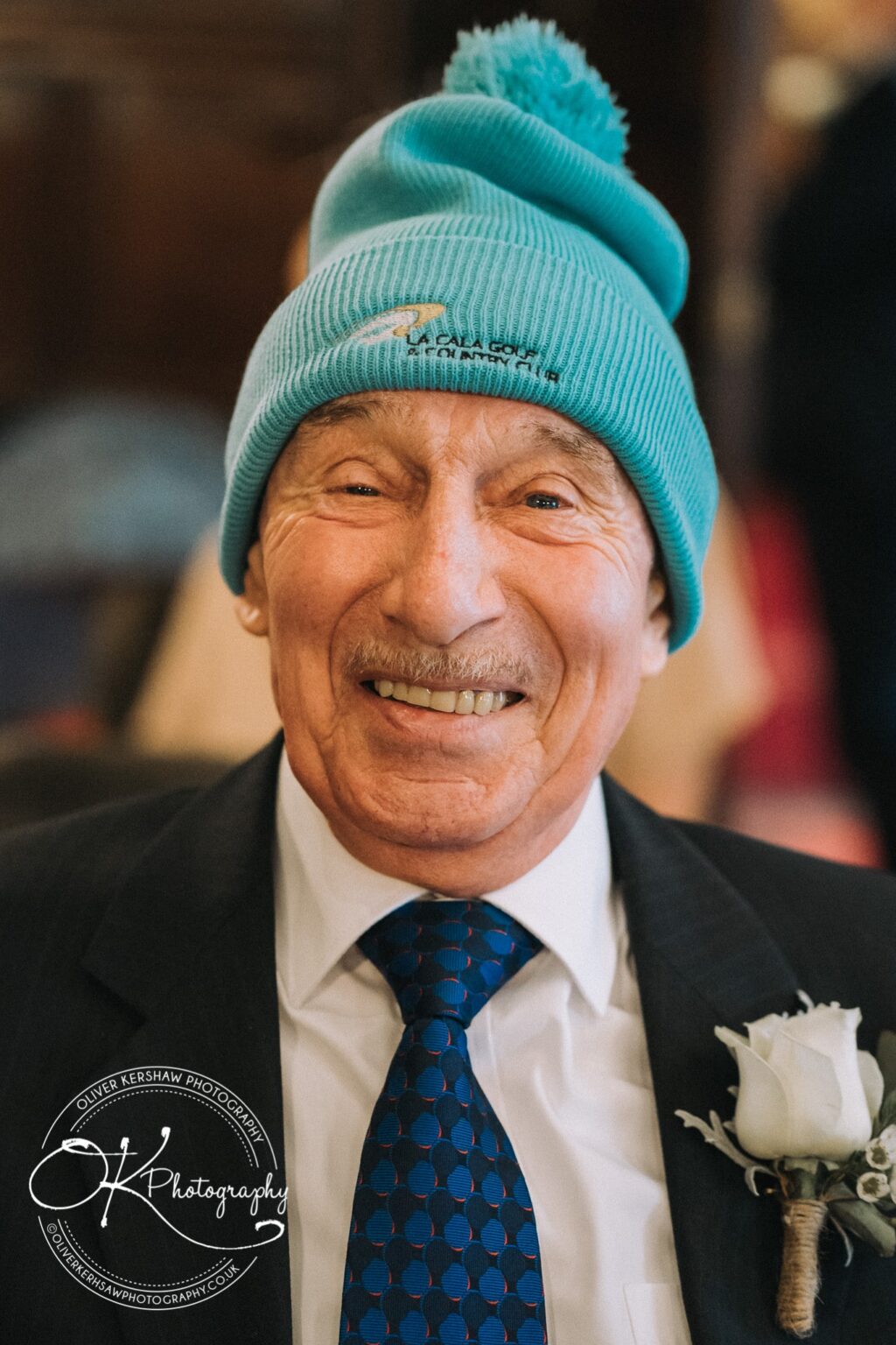 Wedding Photography George Hotel Stamford Smiling elderly man in a suit and tie wearing a bright blue beanie hat with a white flower boutonnière.