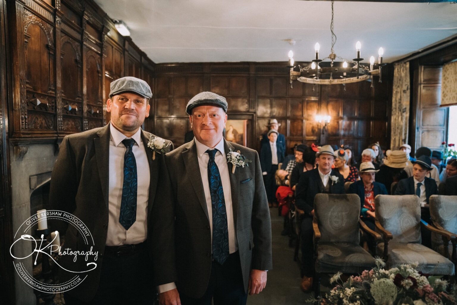 Wedding Photography George Hotel Stamford Two men wearing suits and flat caps standing in a wood-panelled room with seated guests in the background.