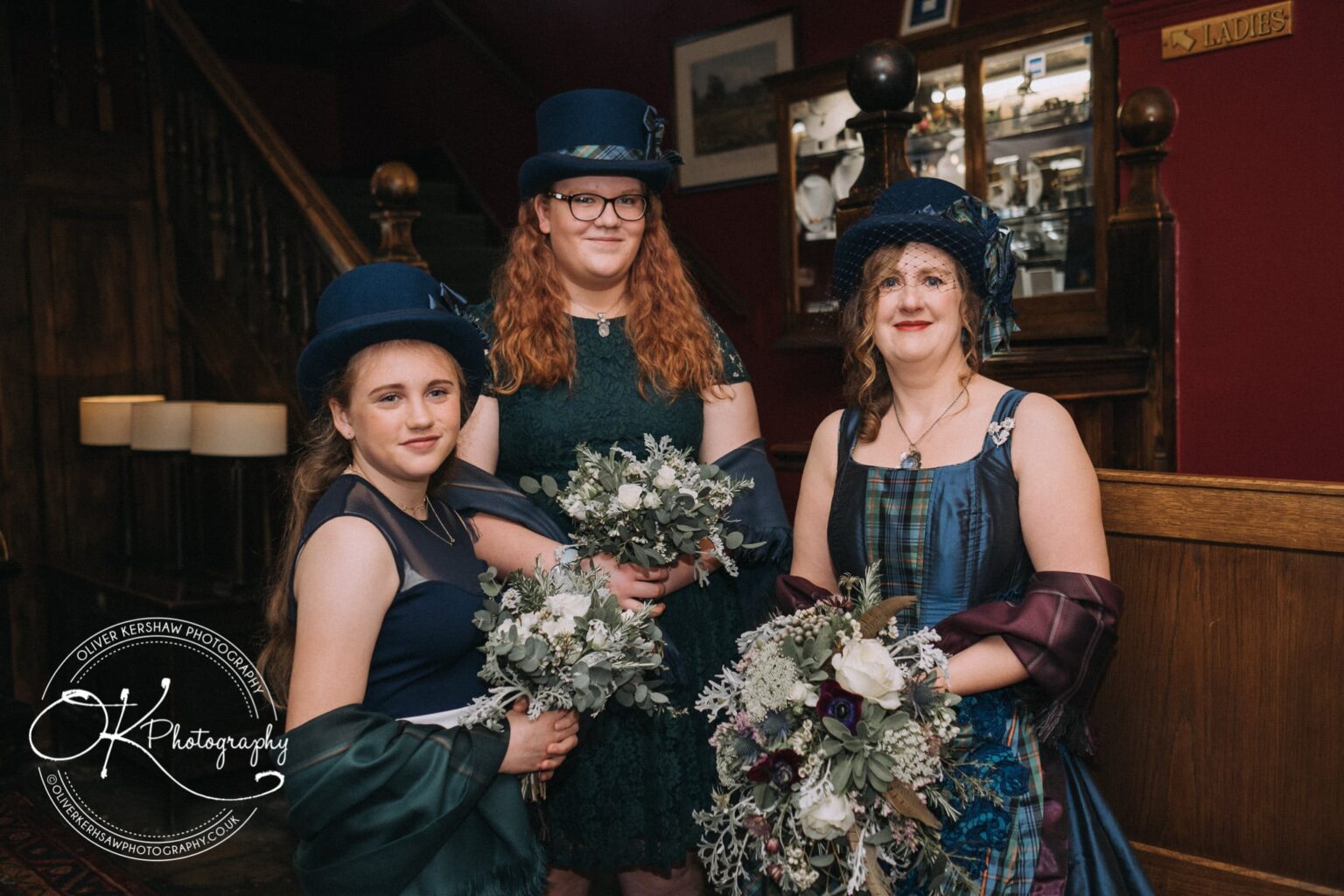 Wedding Photography George Hotel Stamford Three women in formal attire and blue hats posing with bouquets inside a vintage-style hotel with wooden décor.