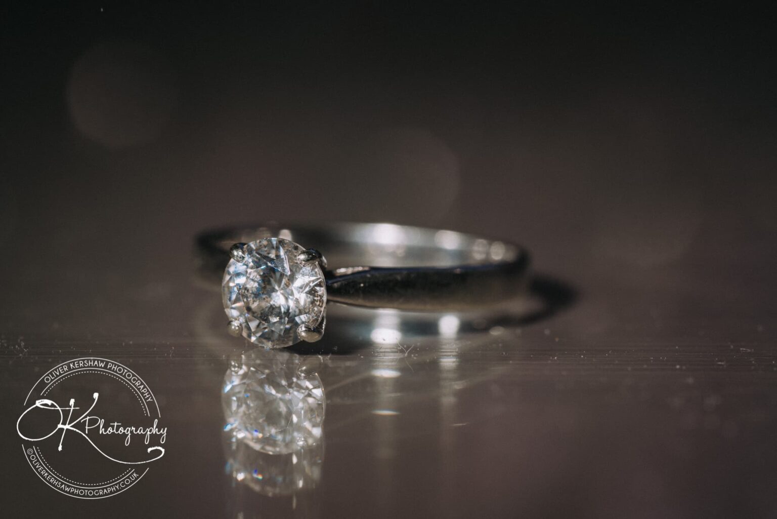 Wedding Photography George Hotel Stamford A close-up image of a diamond engagement ring with a reflection on a shiny surface, featuring a logo for "Oliver Kershaw Photography" in the bottom left corner.