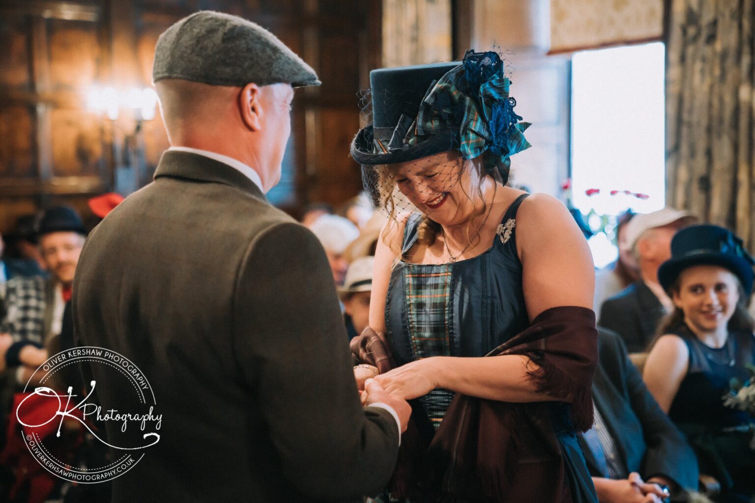 Wedding Photography George Hotel Stamford A couple exchanging rings during a ceremony, with the woman wearing a decorative hat and the man in a suit, in a warmly lit room with onlookers.
