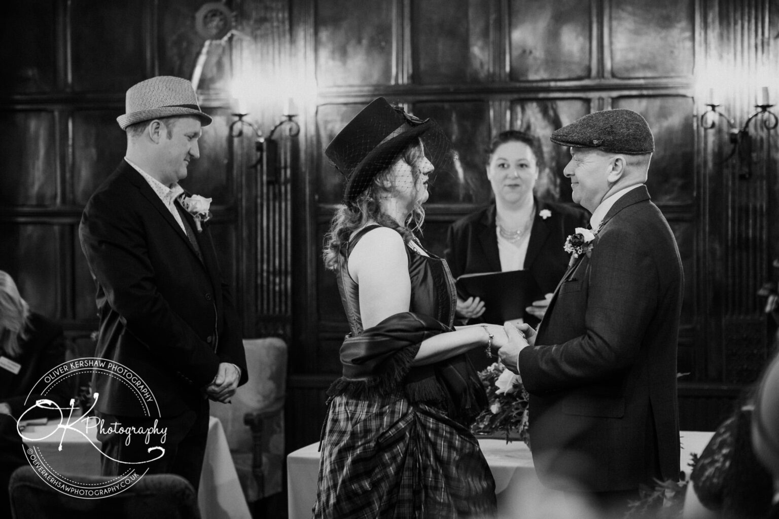 Wedding Photography George Hotel Stamford A black and white photo of a wedding ceremony in a wood-panelled room, with a couple holding hands while a celebrant officiates. Two other individuals stand nearby, all dressed in formal attire.