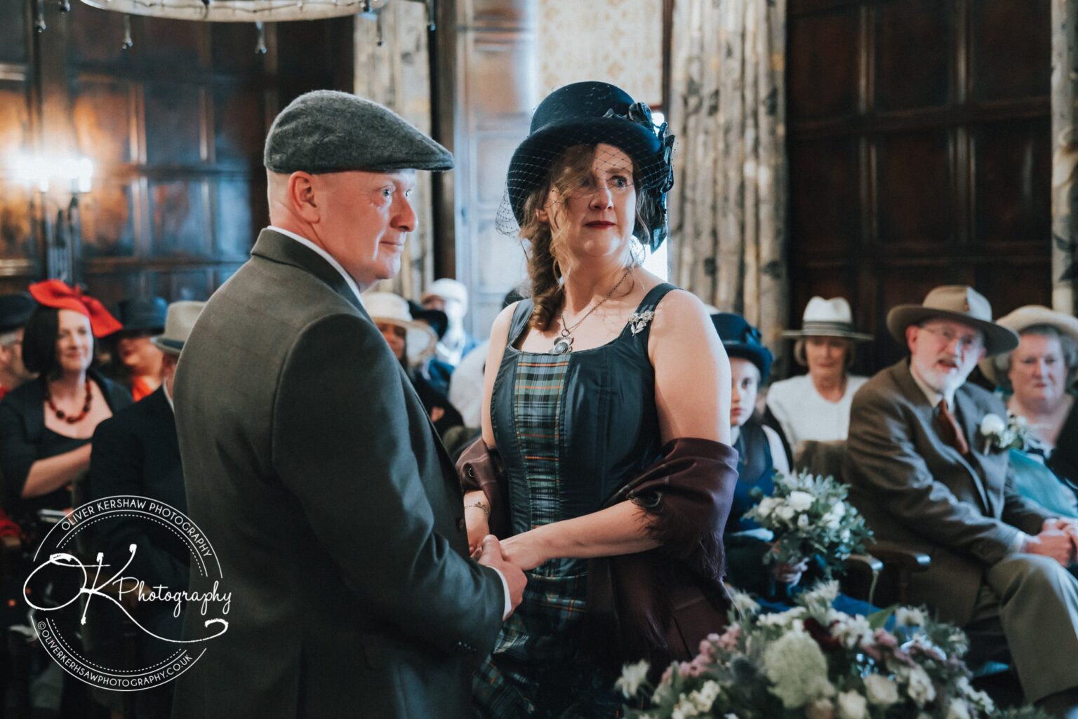 Wedding Photography George Hotel Stamford A couple dressed in formal attire holding hands, surrounded by seated guests wearing hats, inside George Hotel Stamford.