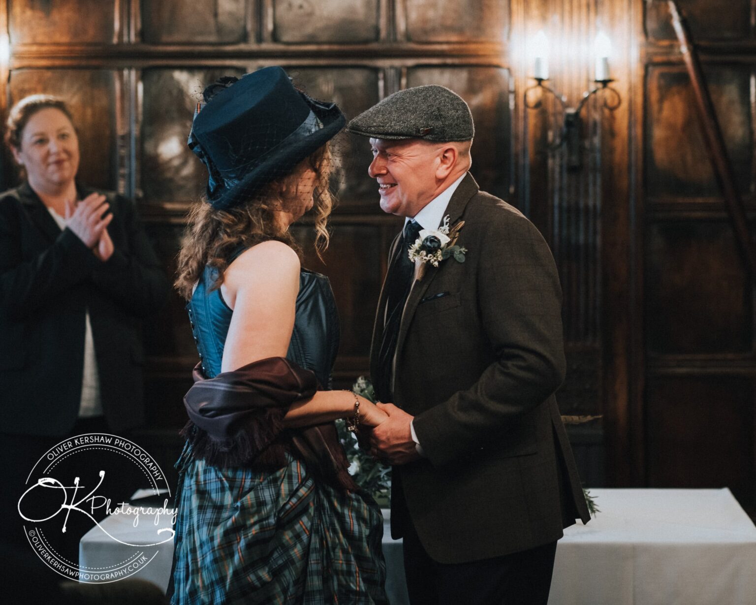 Wedding Photography George Hotel Stamford A couple in formal attire smiling at each other while a woman claps in the background, inside a wood-panelled room.