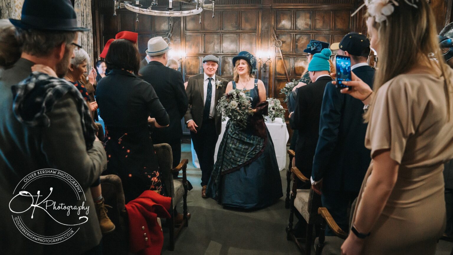 Wedding Photography George Hotel Stamford A bride and groom in formal wear walking down the aisle in a warmly lit, wood-panelled room filled with seated guests, some of whom are capturing the moment on their phones.