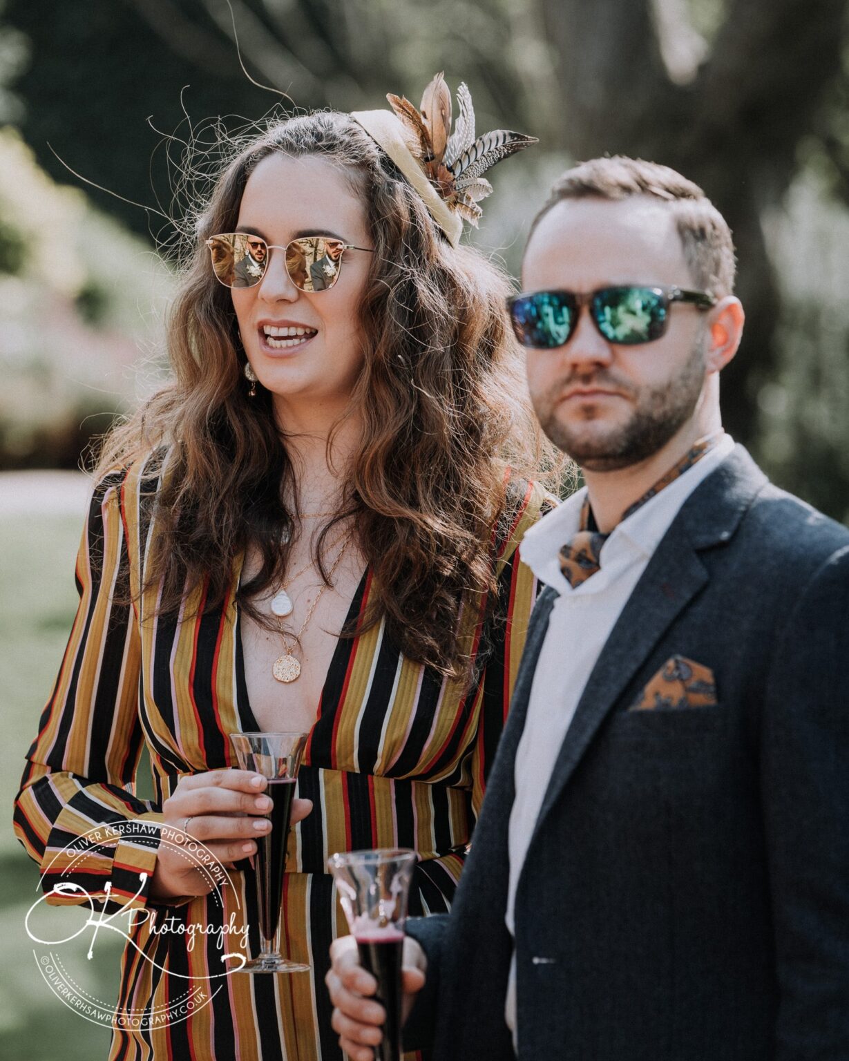 Wedding Photography George Hotel Stamford A man and a woman wearing sunglasses and holding drinks at an outdoor event, with "Olliver Kershaw Photography" watermark on the image.