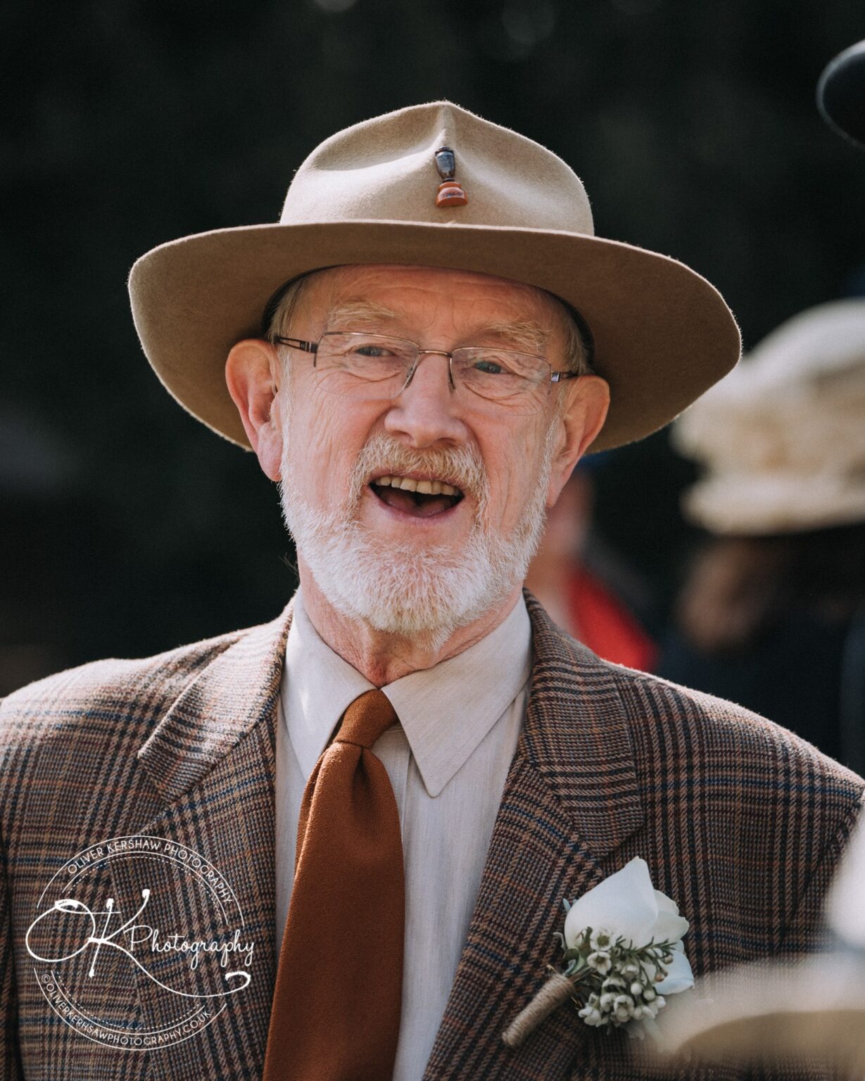 Wedding Photography George Hotel Stamford An elderly man with a white beard, wearing a wide-brimmed hat, glasses, a checked blazer, and a brown tie, smiling.