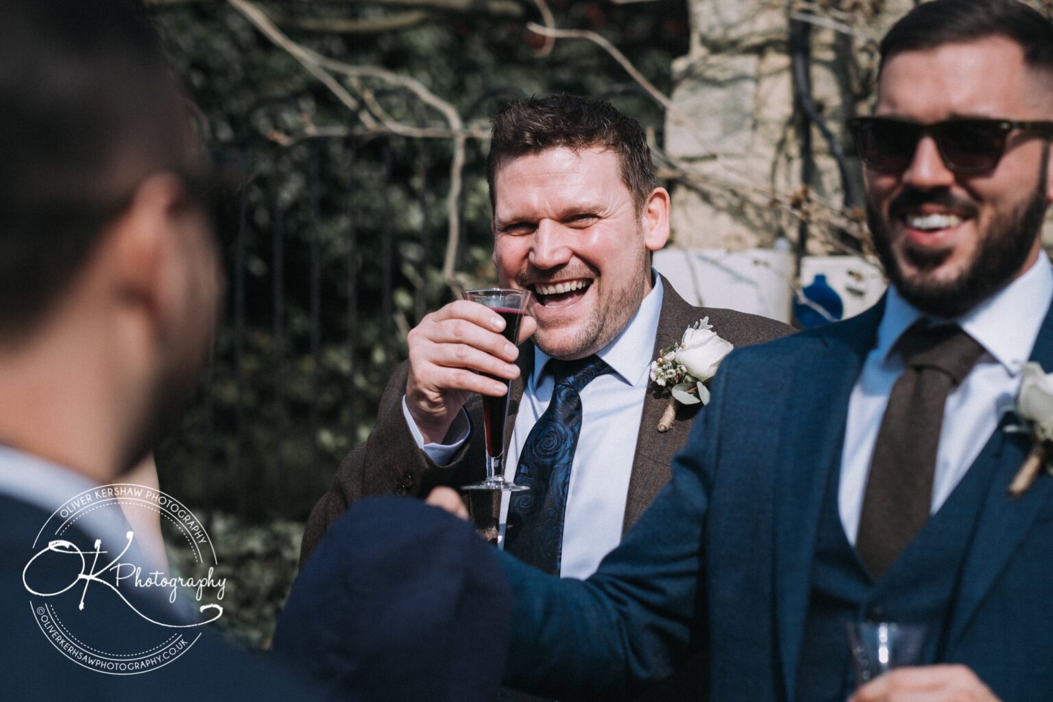 Wedding Photography George Hotel Stamford A man in a suit holding a glass and laughing with others at an outdoor event.