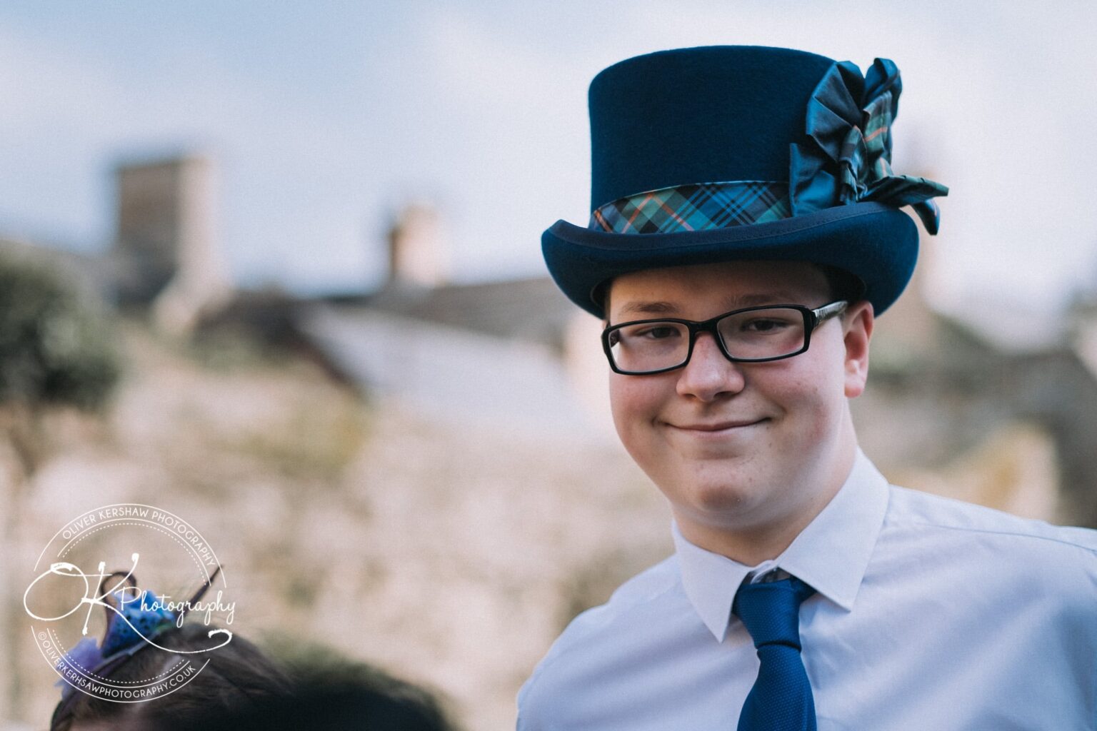 Wedding Photography George Hotel Stamford A young person wearing glasses, a white shirt, blue tie, and a blue top hat with a tartan ribbon, smiling in an outdoor setting.
