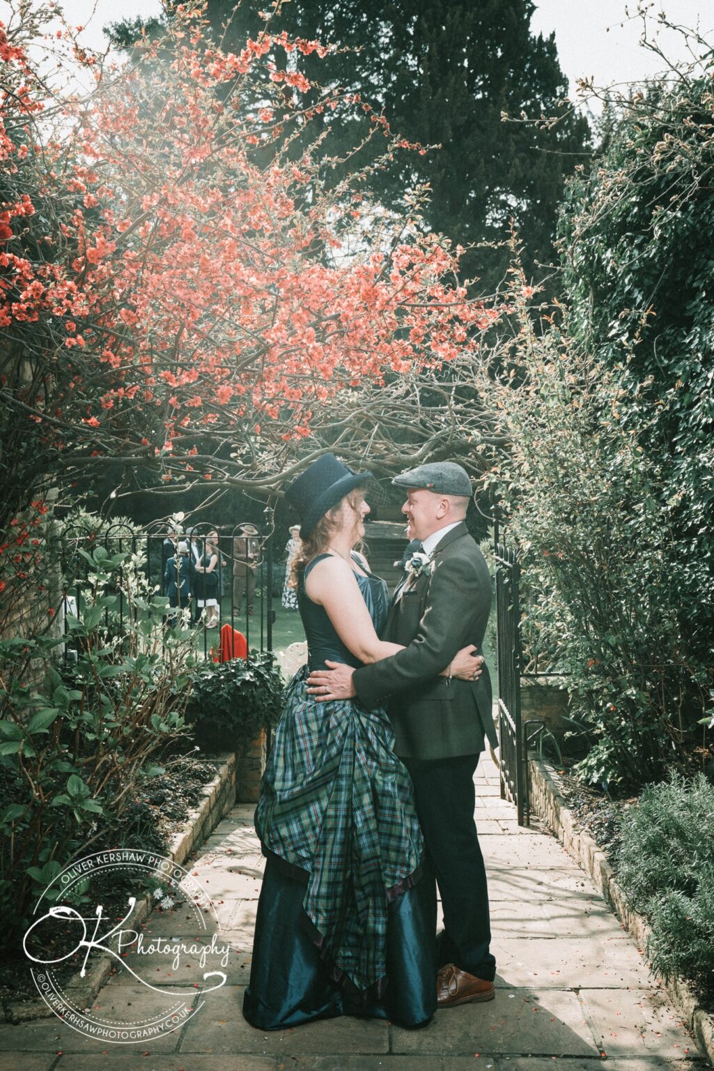 Wedding Photography George Hotel Stamford A couple in formal attire embraces on a garden pathway, surrounded by lush greenery and a flowering red bush.