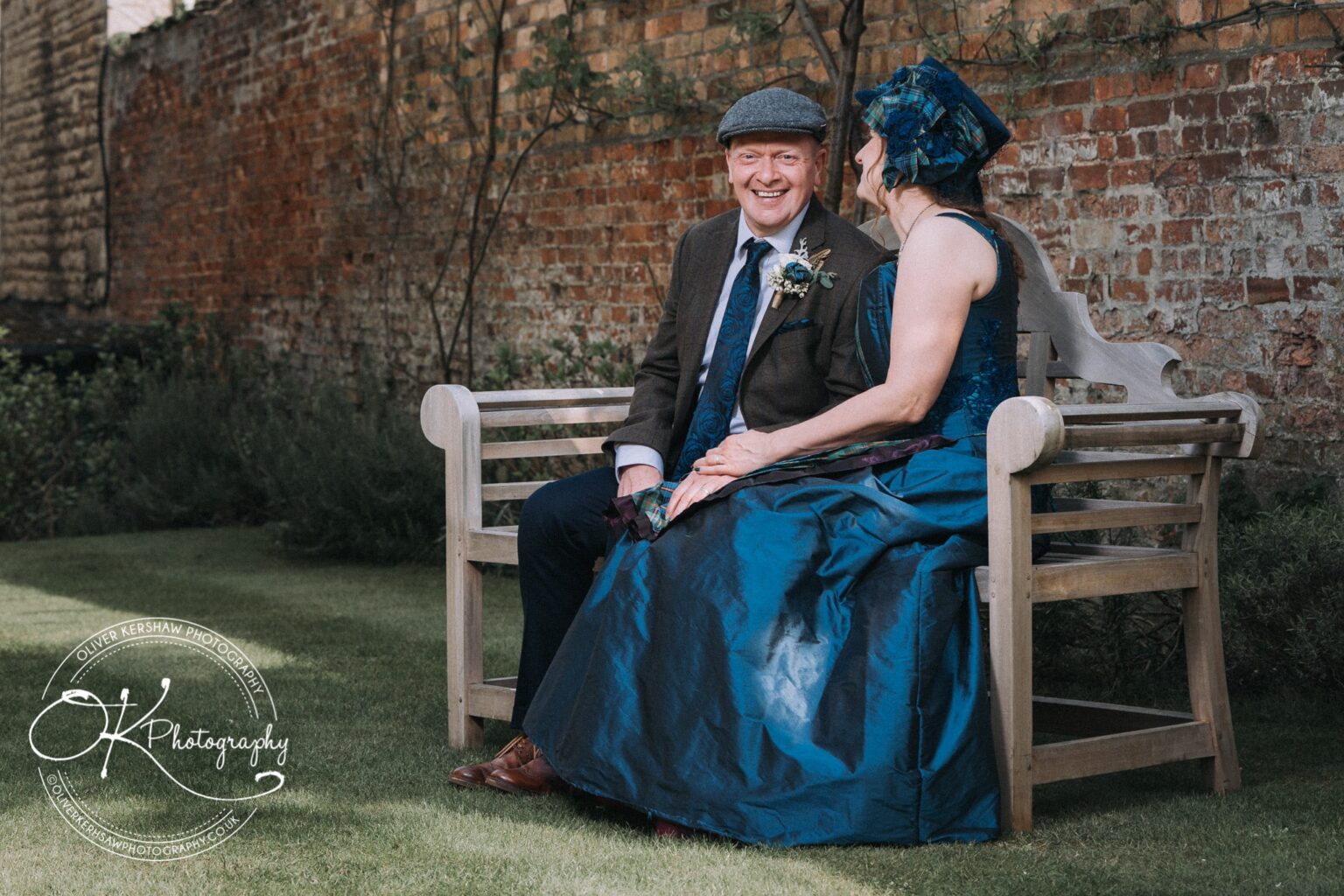 Wedding Photography George Hotel Stamford A smiling man and woman in formal attire sitting on a wooden bench in a garden with a brick wall background.
