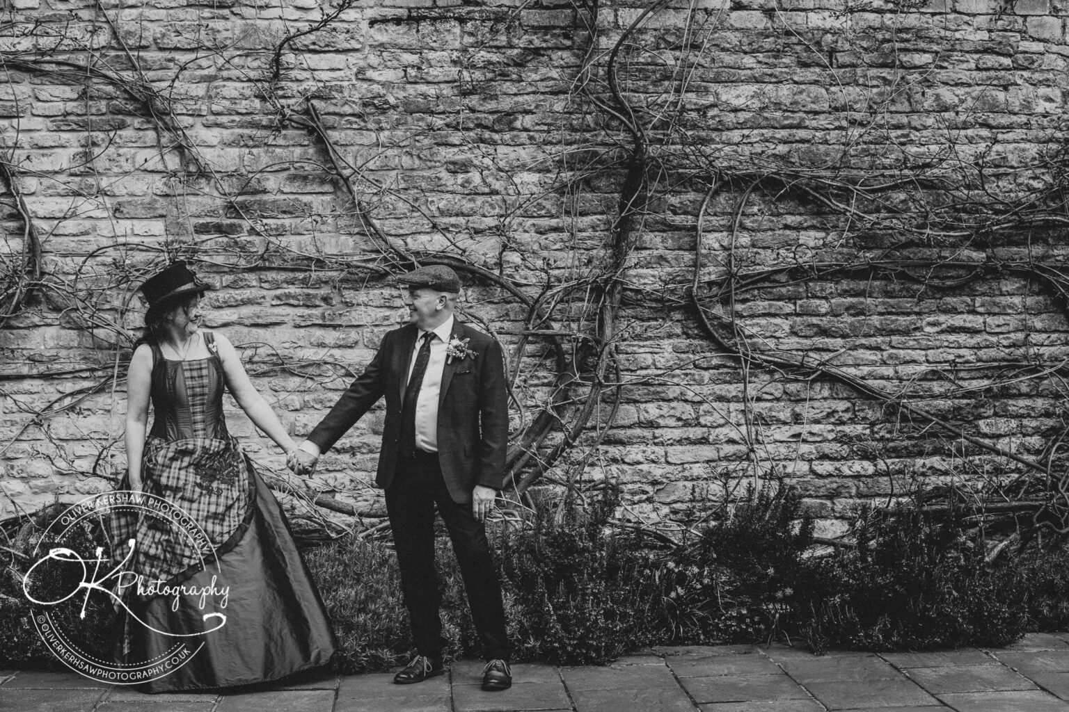 Wedding Photography George Hotel Stamford A couple holding hands and looking at each other, dressed in formal attire including a suit and top hat, standing against a stone wall with vines.