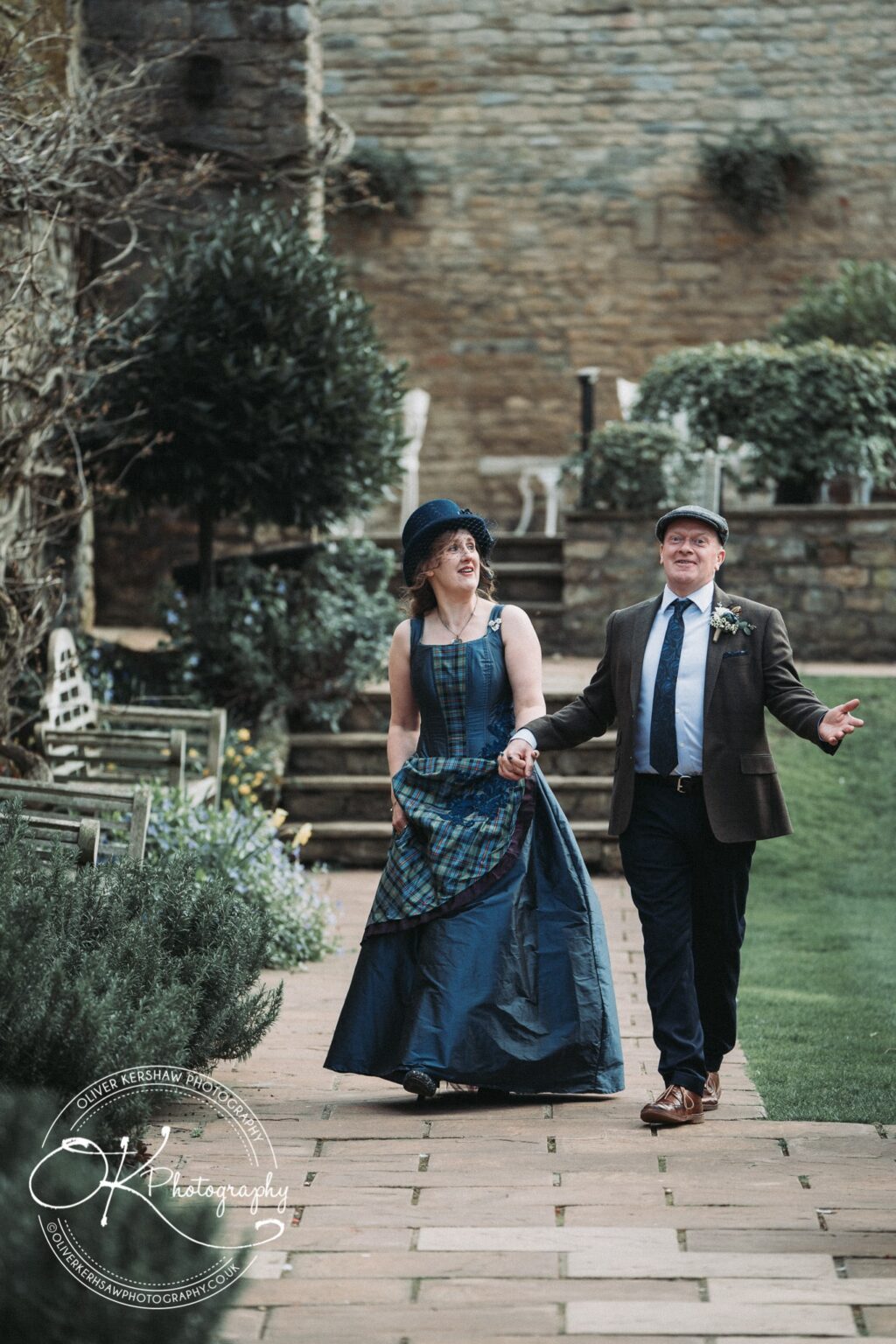 Wedding Photography George Hotel Stamford Two people walking and holding hands, dressed in vintage attire, in an outdoor garden setting with a stone wall and greenery in the background.