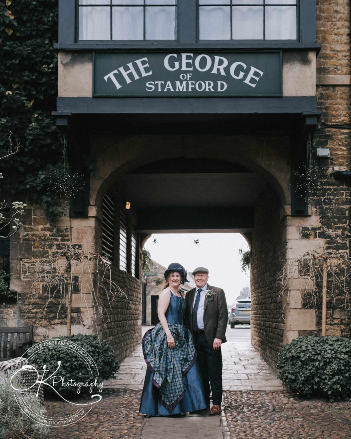 Wedding Photography George Hotel Stamford A couple dressed in formal attire stand smiling under the archway entrance of The George of Stamford hotel.
