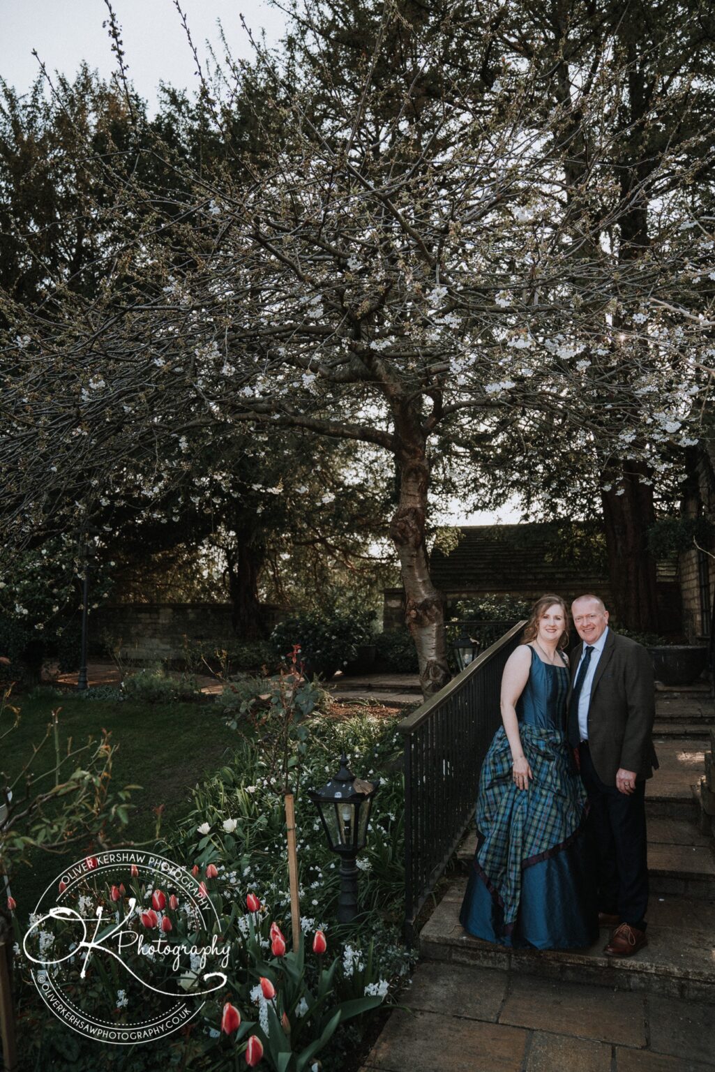 Wedding Photography George Hotel Stamford A couple posing together beneath a tree in a garden, with flower beds and lamp posts in the background.