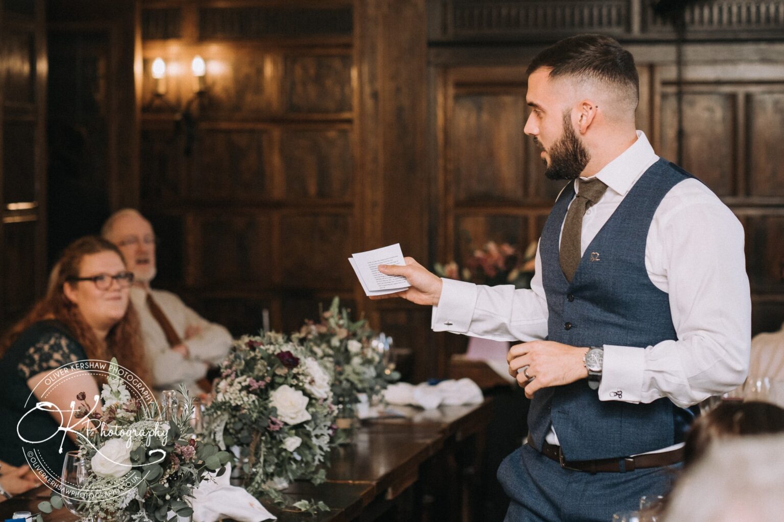 Wedding Photography George Hotel Stamford A man in a blue vest speaks to seated guests, holding a piece of paper, with floral arrangements and wooden panelling in the background.
