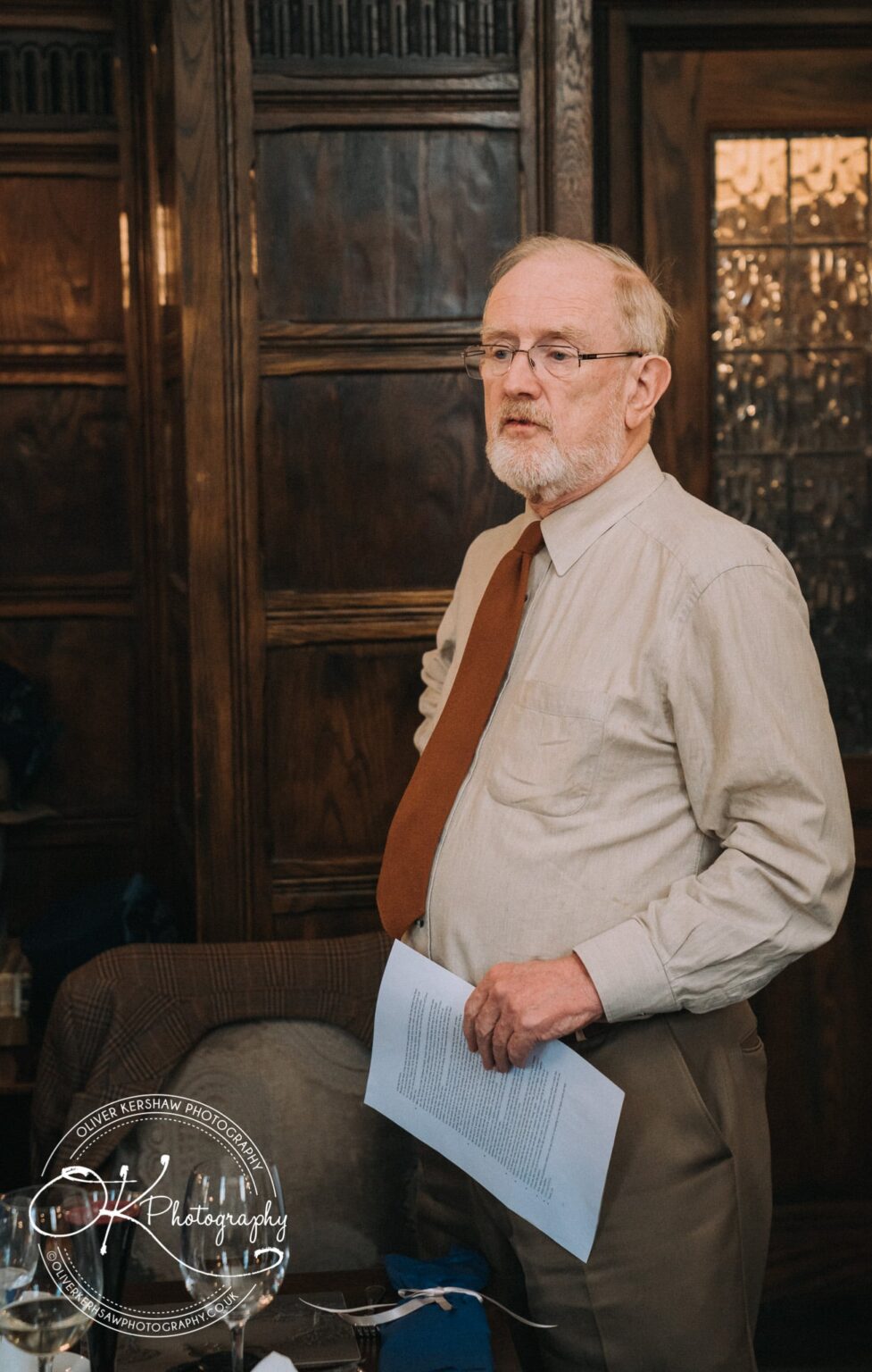 Wedding Photography George Hotel Stamford An older man with a white beard and glasses, wearing a beige shirt and brown tie, stands holding a piece of paper in a wood-panelled room.
