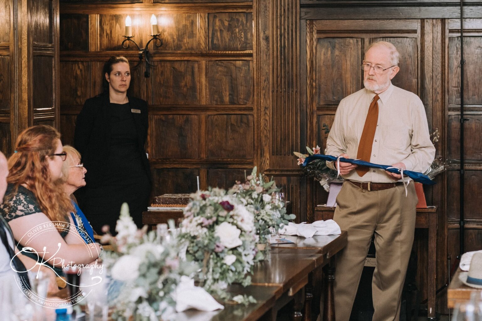 Wedding Photography George Hotel Stamford A man standing and speaking while holding a blue folder in a wood-panelled room, with a woman standing in the background and several people seated at tables adorned with flowers.