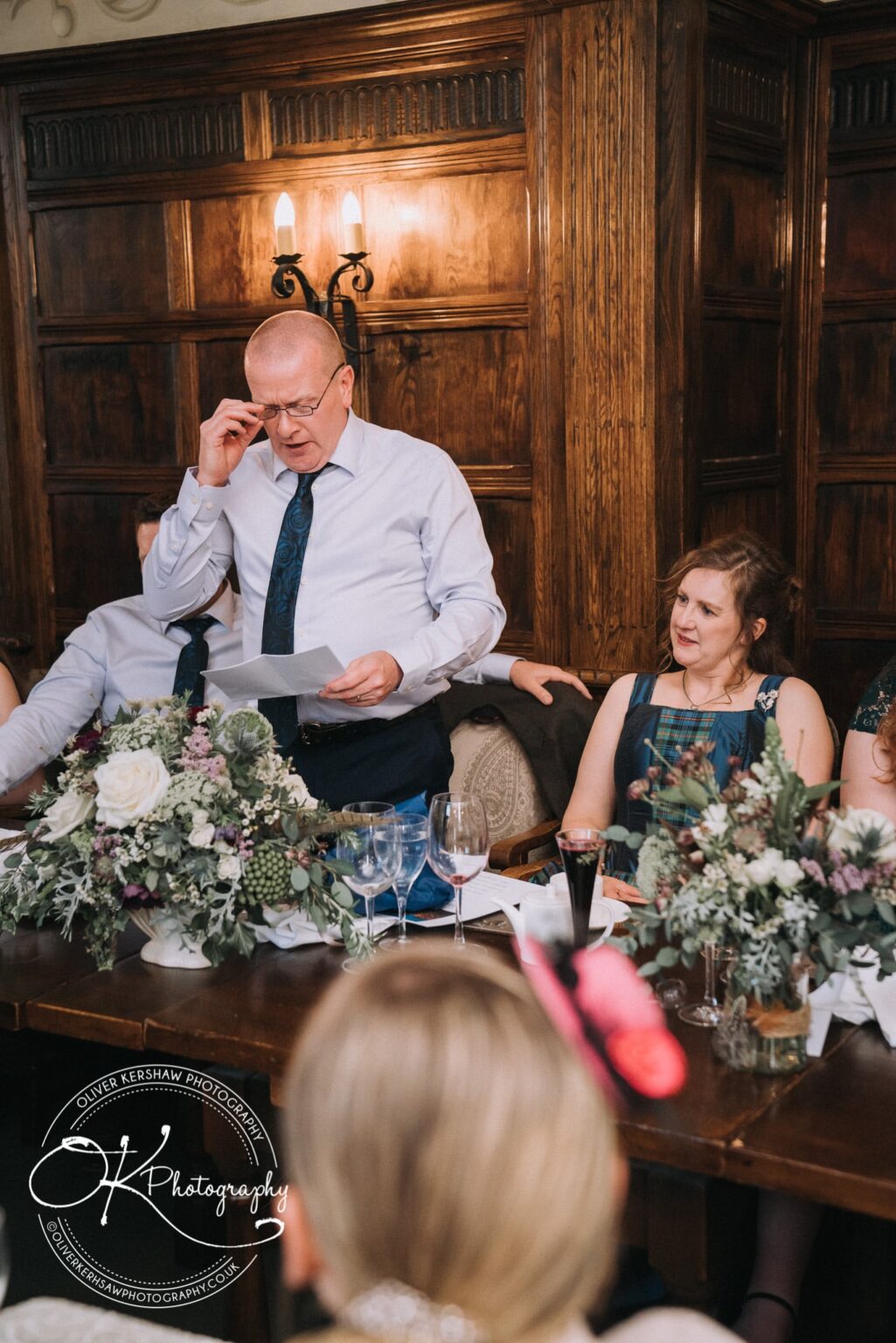 Wedding Photography George Hotel Stamford A man giving a speech inside a wood-panelled room, with floral arrangements and wine glasses on the table, and people seated around him.